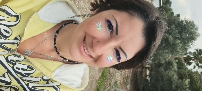 A close-up, waist-up portrait captures an adult female smiling and looking directly at the camera, located outdoors in the Mate Asher Regional Council, Israel. The subject has short, dark hair, prominent blue eyeliner on her upper and lower eyelids, and iridescent glitter applied in snowflake-like patterns below each eye. She features a small stud piercing in her left nostril and a hoop piercing in her right nostril. Her left ear displays a dangling earring with multiple chains and a decorative antler-like element. Around her neck are two necklaces: an upper choker-style necklace composed of black beads with silver spacers, and a lower, longer silver chain featuring a round "Tree of Life" pendant. Fine glitter is visible on her upper chest and collarbone area. She is dressed in a yellow hooded sweatshirt with white lining and a partially visible black word, likely "Detroit," printed on the front. The background consists of numerous olive trees with dark green foliage, a tall palm tree in the upper right, and dry, light-colored ground. A white structure is partially visible on the far left. The sky is overcast.