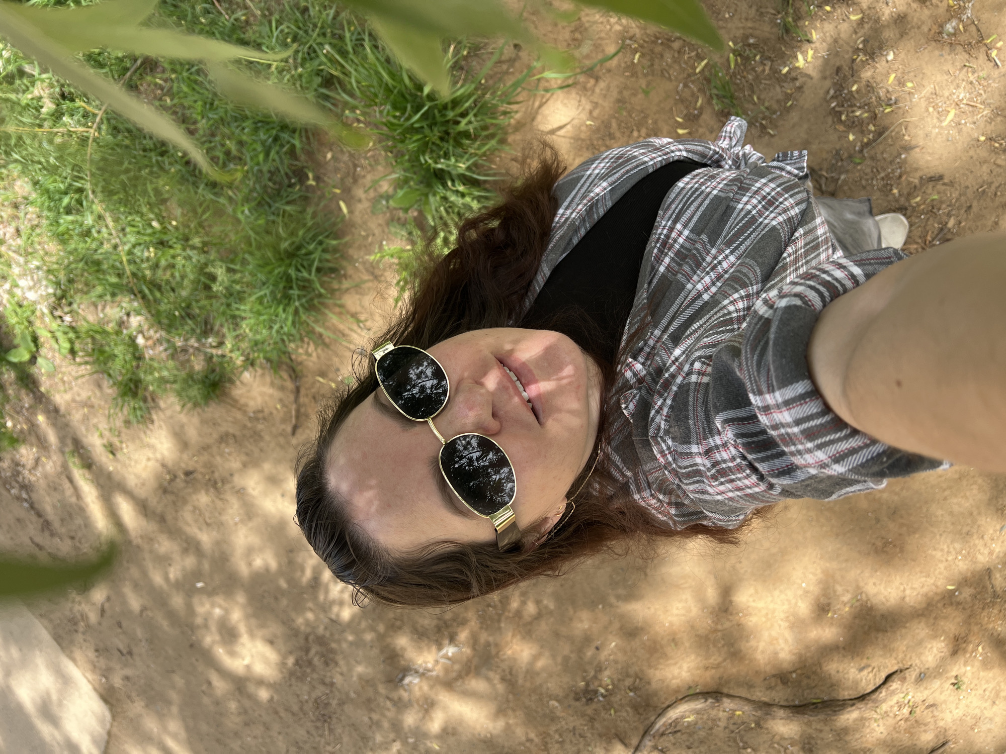 This media file captures a high-angle view of a person outdoors in Noviy, Russia. The subject, who appears to be a woman, is looking upwards towards the camera with a slight smile, revealing a hint of her teeth. She has long dark hair and is wearing gold-rimmed round sunglasses that reflect the surrounding foliage. Her attire consists of a black top layered under a grey, white, and red plaid shirt, which is tied at the front. A delicate gold chain is visible around her neck.
The scene is an outdoor setting with a mix of light brown dirt or sand and patches of green grass on the ground. Blurry green foliage, likely from an overhead tree, is prominent in the foreground and casts dappled shadows across the ground and on the person's face and body. The bright, clear shadows indicate that it is daytime and sunny. There are no visible signs of text or specific events occurring beyond the portrait itself. The person's expression suggests contentment or casual posing for the camera.