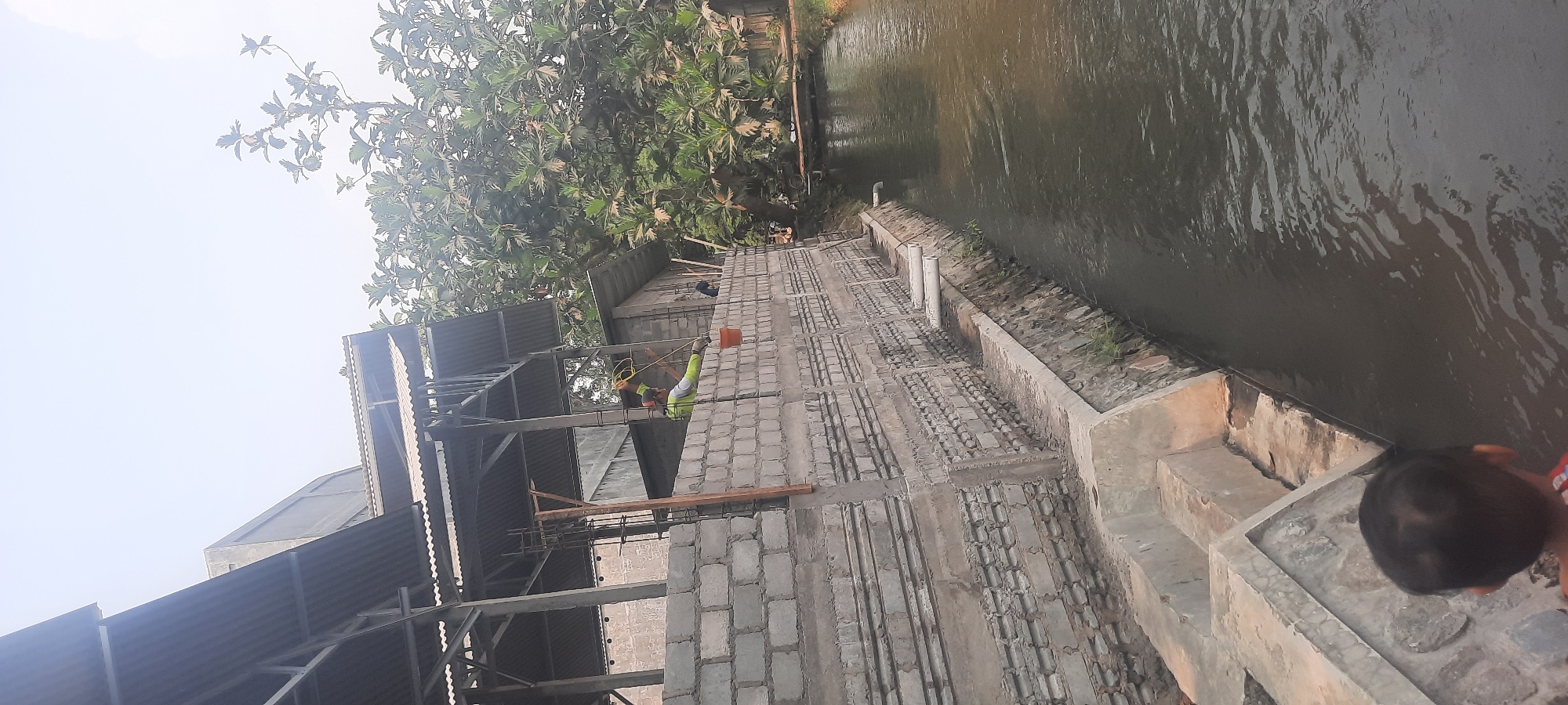 This image shows a construction site next to a body of water, likely a canal or small river. The construction appears to be of a building with a corrugated metal roof supported by metal beams and concrete pillars. The walls are made of concrete blocks. Two construction workers are visible, one in a lime green shirt is holding yellow wires and appears to be working on the structure. An orange bucket is hanging from the wall.
In the foreground, a young child's head is visible, looking towards the water. The water is dark and reflects the sky and surrounding greenery. A large tree with broad leaves is prominent on the right side of the image, its branches extending towards the construction. The environment suggests an outdoor setting in Ploso, Indonesia, likely a developing area where construction is taking place near a waterway.
