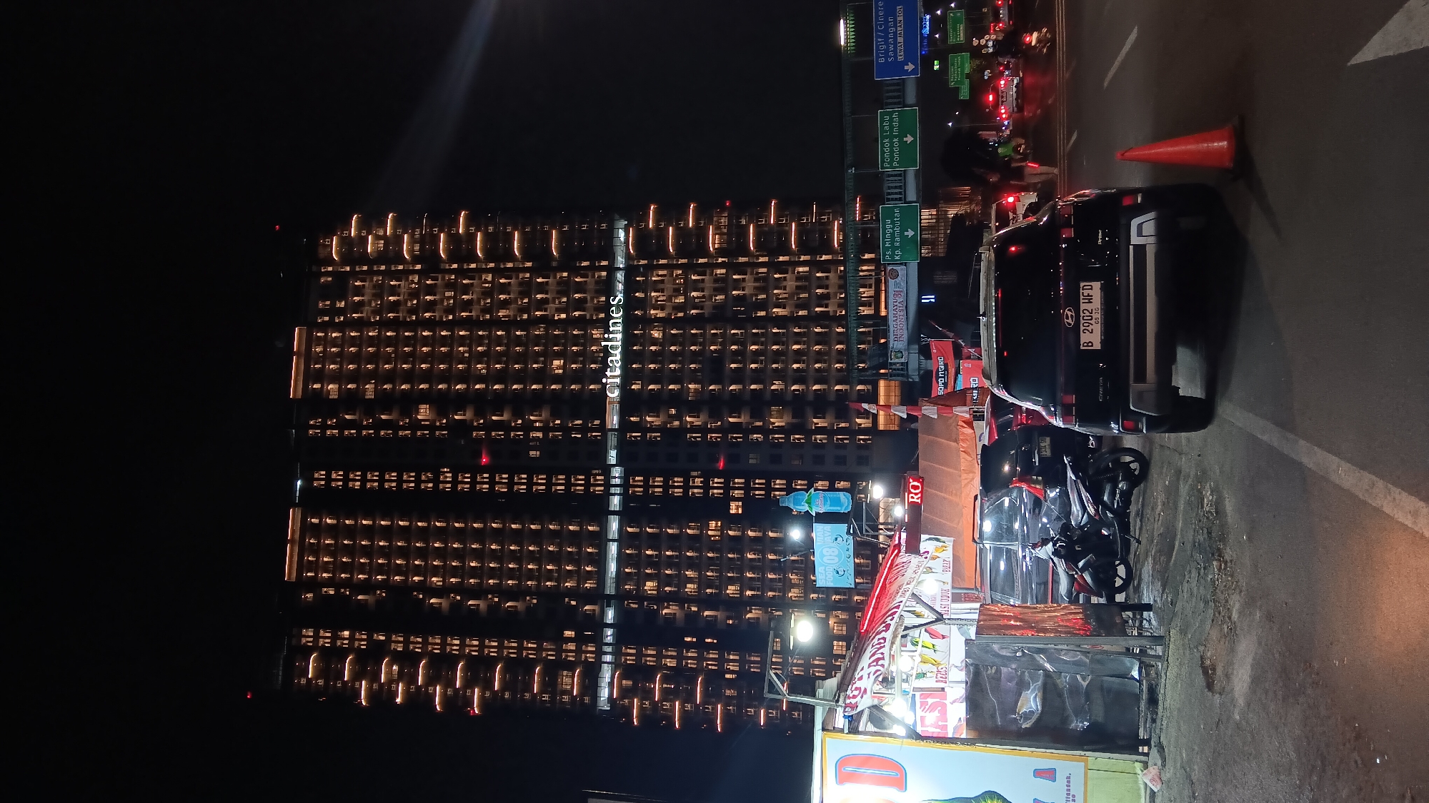 The image shows a street scene at night in South Jakarta, Indonesia, dominated by a tall, illuminated apartment building with the "citadines" logo visible. In the foreground, there are several vehicles, including a black Hyundai Creta with license plate B 2902 HFD, a motorcycle, and other cars. A street food stall with red and white awnings is set up on the left, displaying various food items. Traffic signs for Pondok Labu, Pondok Indah, and Brigif/Cinere are visible on an elevated road structure in the background. The sky is dark, and the scene is lit by the building's lights, streetlights, and vehicle headlights.