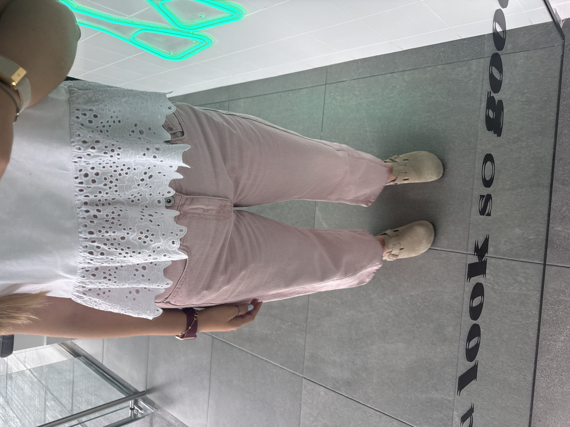 The image provides a partial, overhead view of a person standing indoors. The person is wearing a white top with intricate eyelet lace or broderie anglaise detailing and light pink (or dusty rose) straight-leg jeans or pants. Their feet are clad in light tan or beige closed-toe flat shoes, resembling clogs or slip-ons. On the person's left wrist, a maroon-strapped watch and a gold-toned bracelet are visible, along with a ring on one finger.
The setting is an indoor space with modern grey tiled flooring and white tiled walls. A bright green or turquoise neon light fixture with an angular, abstract design is visible on the white ceiling. To the right of the person, a clear glass barrier or display surface is present, reflecting parts of the floor.
Prominently displayed on the grey floor, and also reflected in the glass barrier, are large black block letters that spell out "LOOK SO GOOD". The overall scene suggests a casual moment, likely within a retail environment, a modern public building, or a gallery in San Bernardino Tlaxcalancingo, Mexico. The lighting is artificial, and no specific time of day or outdoor weather conditions are discernible. The person appears to be standing or casually walking through the space.
