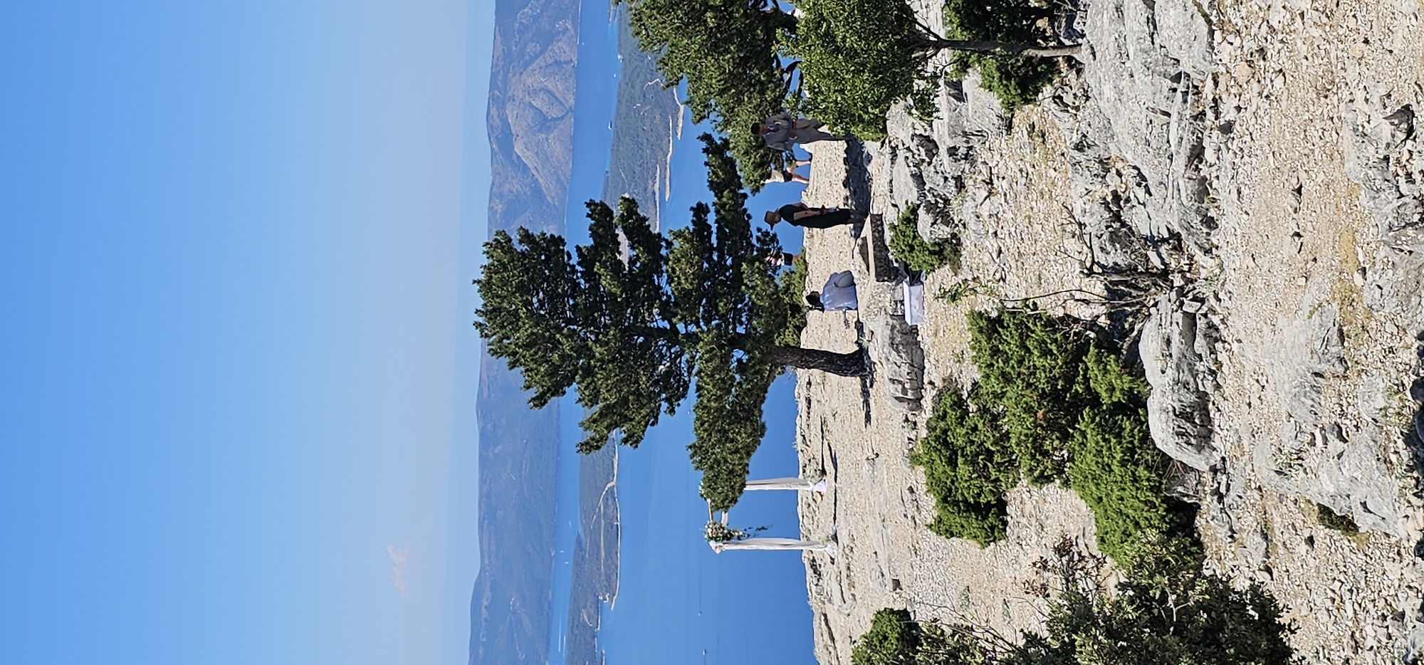 This is a vertical, eye-level, full shot of a wedding setup on a rocky clifftop overlooking a body of water and islands. The sky is clear and blue. In the foreground, there is a wedding arch draped with white fabric and adorned with flowers. A large evergreen tree stands to the right of the arch, with its branches extending over the rocky terrain. Several people are visible in the midground, some standing and some sitting. One person in a white shirt sits to the left of the tree, facing away from the camera. Another person in a dark outfit and a hat stands near the tree, facing towards the body of water. To the right of them, two more people are visible, one in a suit and one in a dress. A white box is placed on the ground near the seated individual. The clifftop is covered in rocks and sparse green vegetation. In the background, a vast expanse of blue water is dotted with small islands and boats. The overall scene suggests an outdoor wedding ceremony in a scenic, natural setting. The location appears to be the Vidova Gora peak on the island of Brač, in Općina Bol, Croatia, known for its panoramic views of the Adriatic Sea and surrounding islands.