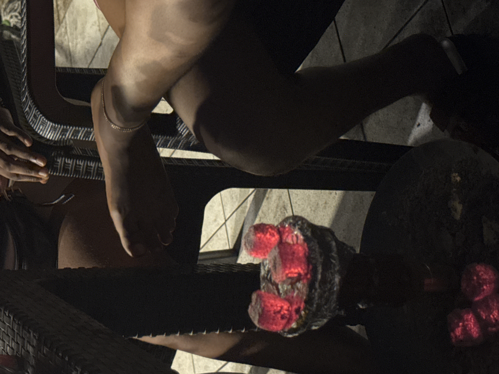 A person is sitting on a black wicker chair outdoors. Their legs are crossed, and they are wearing an anklet. In the foreground, there is a hookah with glowing coals. The location appears to be in Rumuomasi, Nigeria. The environment is dark, suggesting it is night.