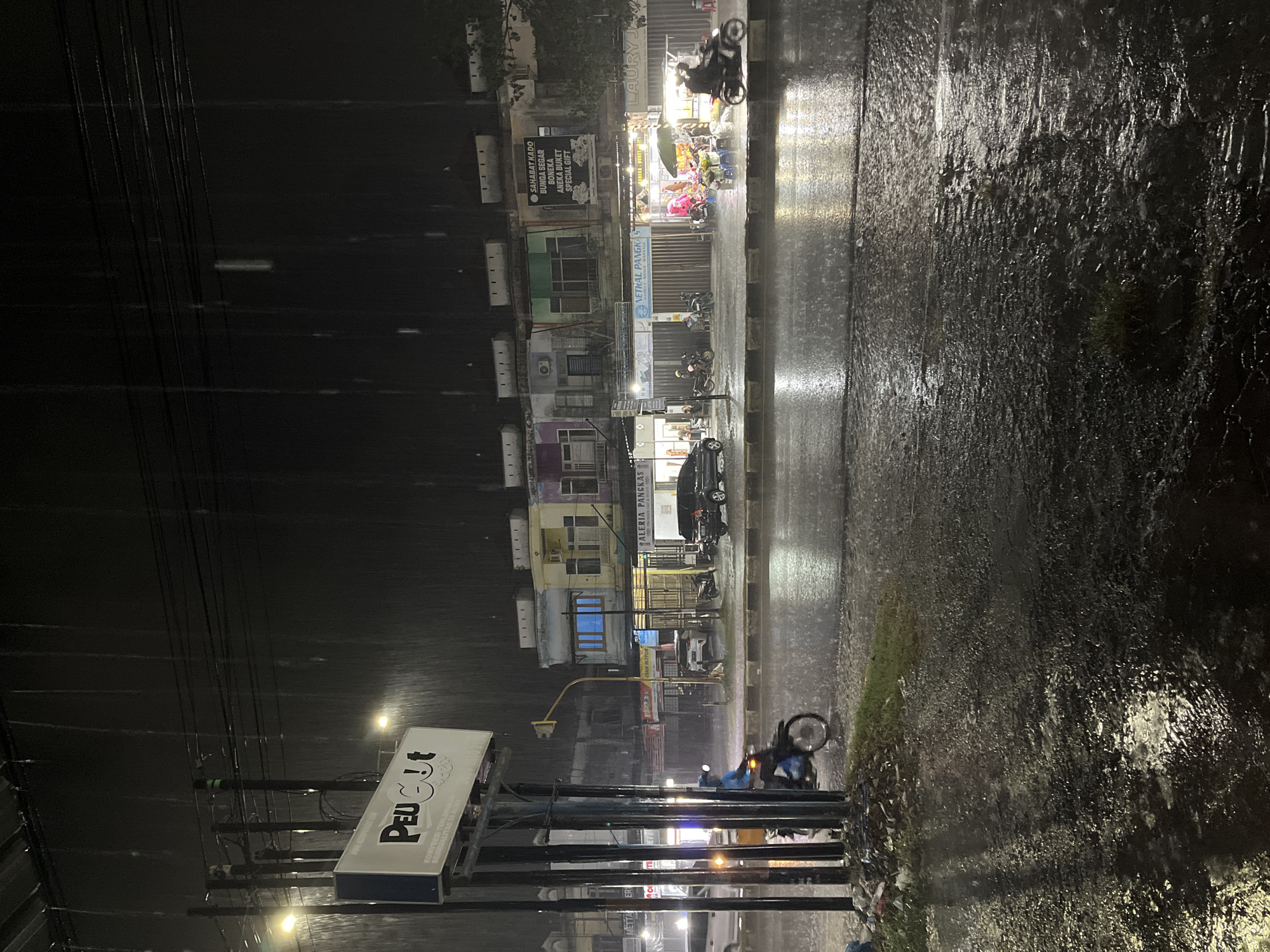 This is a medium shot of a street scene in Banda Aceh, Indonesia, at night during a heavy rain. The foreground is dominated by a wet, reflective asphalt road, with puddles and streaks of water visible. To the left, a motorcycle with a rider in a blue jacket is moving away from the camera, splashing water. In the mid-ground, a row of commercial buildings lines the opposite side of the street. These buildings are illuminated by streetlights and the interior lights of the shops. One shop, labeled "NETRAL PANGKAS," appears to be a barbershop, and another has a display of flowers. A black car and several motorcycles are parked or in motion on the street. Overhead, power lines are visible against the dark, rainy sky, with streaks of falling rain clearly depicted. The overall atmosphere is dark and wet, characteristic of a tropical downpour.