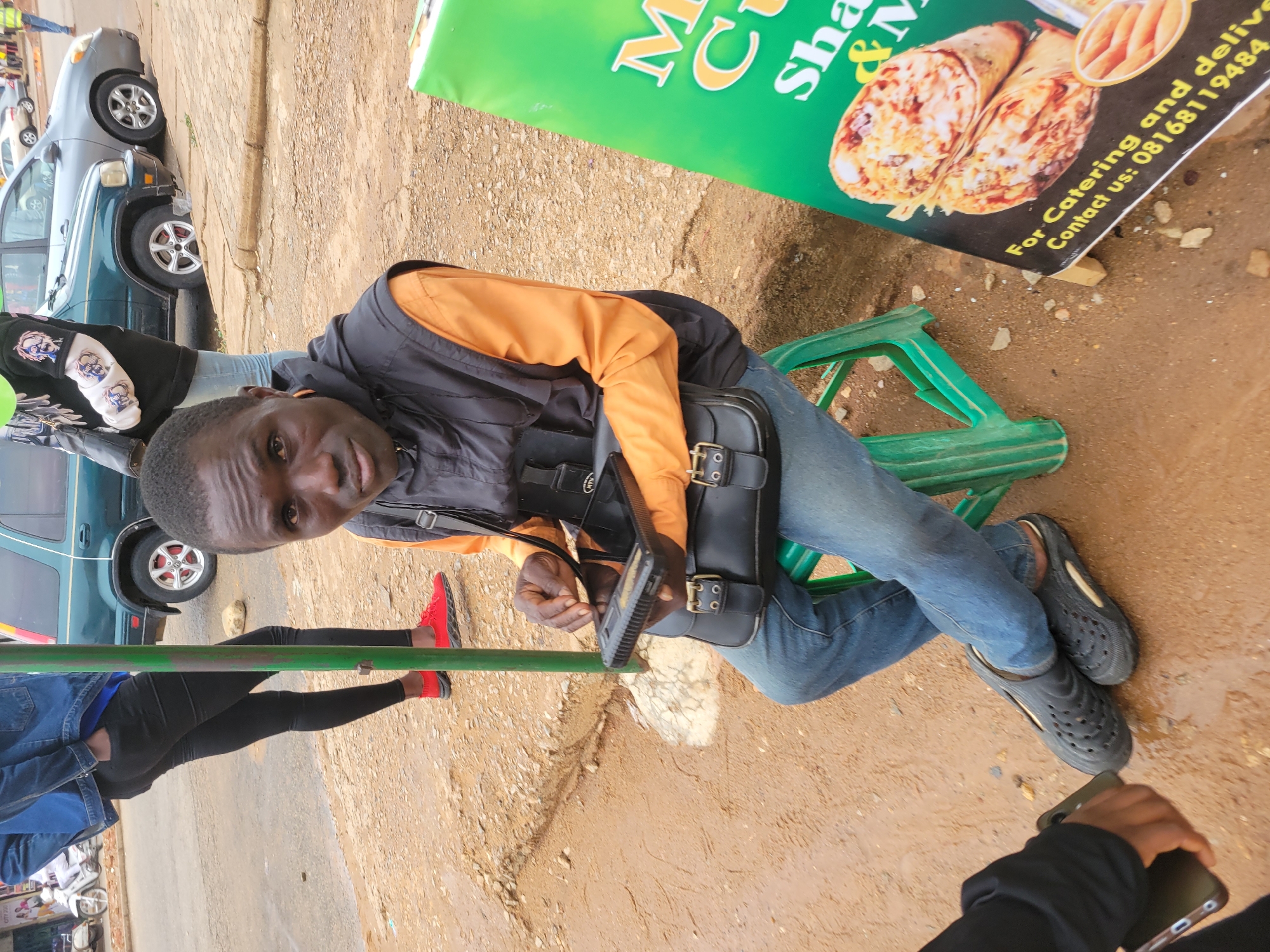 A man sits on a green plastic chair on a dirt surface, wearing blue jeans, a black vest over an orange shirt, and black shoes. He is holding a black bag on his lap and looking at the camera. To his right is a sign that reads "M.C. Snacks & More" with food images and contact information ("For Catering and delivery, Contact us: 08168119484"). To his left, another person wearing red shoes and black pants is standing near a green pole. Behind them, several cars are parked. The location appears to be an outdoor area, likely a roadside or street in Jos, Nigeria.