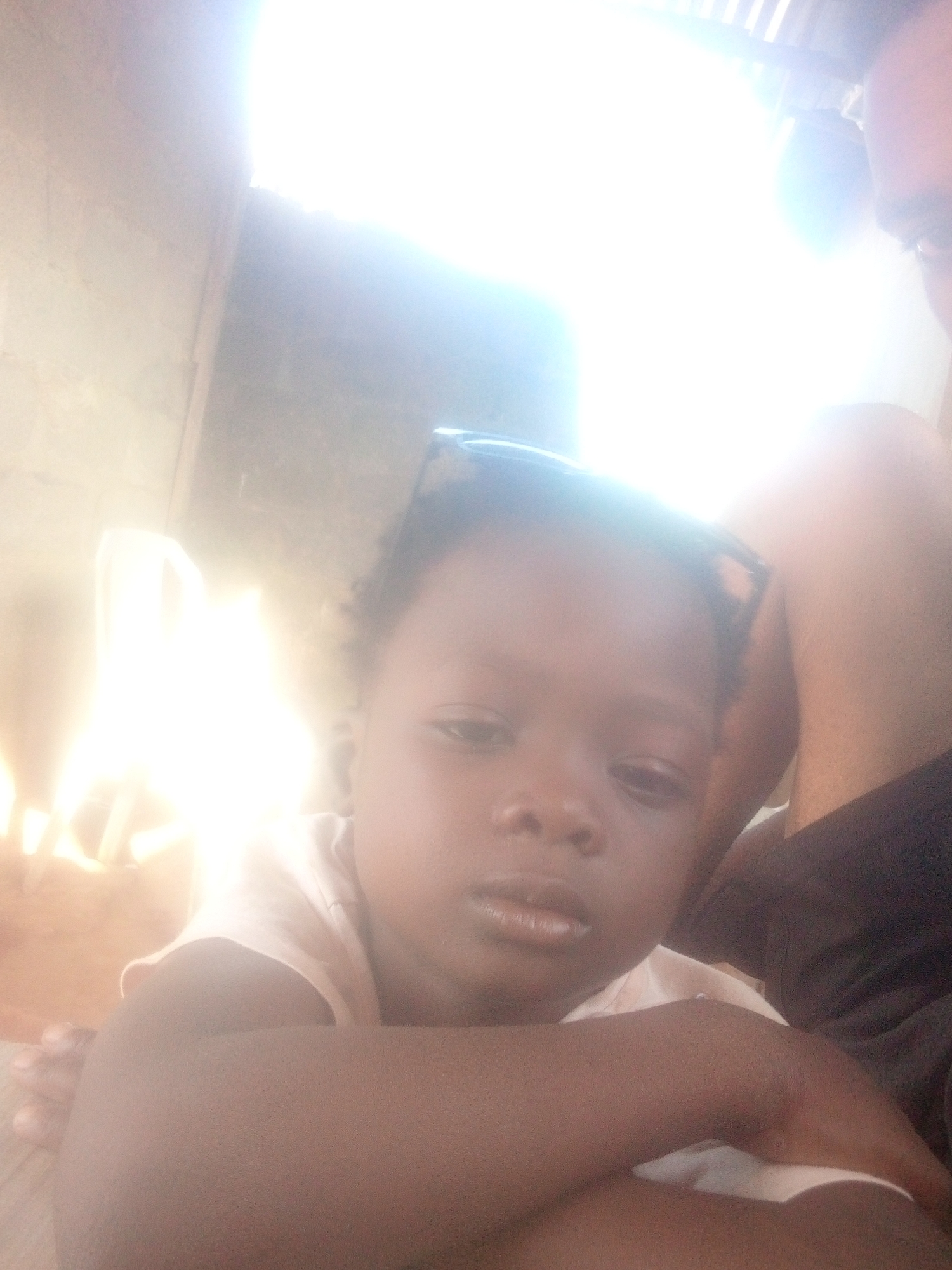 A close-up, slightly overexposed image shows a young child of African descent with sunglasses perched on their forehead. The child has a serious expression. A person's shoulder and leg, likely an adult, are partially visible behind and to the side of the child. The location appears to be indoors, but details are obscured by the overexposure. Location is unknown, Unknown.