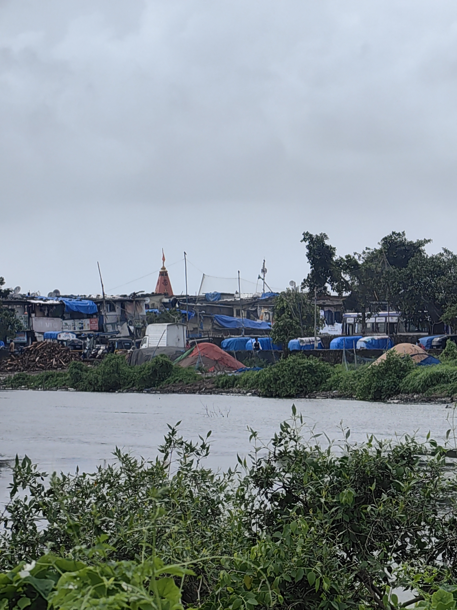 In Mira-Bhayander, India, a waterfront scene unfolds under an overcast sky. The foreground is dominated by lush green foliage, obscuring the lower portion of the view. Beyond the vegetation, a body of calm water stretches across the image. On the opposite bank, a densely populated area is visible, characterized by makeshift structures covered in blue tarpaulins and corrugated metal sheets, indicative of informal housing. A prominent red and orange spire, likely belonging to a temple, rises above the structures. Several vehicles, including a white van and what appear to be auto-rickshaws, are present amongst the buildings. Piles of wood and various materials are scattered throughout the settlement. The overall impression is one of a vibrant, albeit crowded and weathered, urban environment situated by the water.