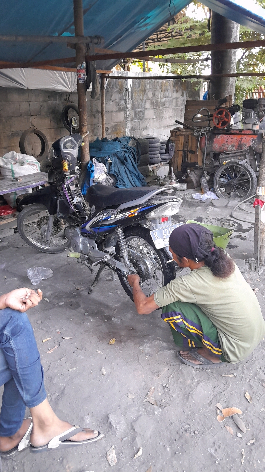 The image depicts a scene in Balun, Indonesia, likely a mechanic's workshop or outdoor repair area. A black motorcycle is parked in the foreground, leaning on its kickstand. A person, dressed in a green shirt and green pants with yellow stripes, is crouched down and appears to be working on the rear wheel or chain of the motorcycle. This person is wearing a dark headscarf and flip-flops. To the left, another person's legs are visible, wearing blue jeans and flip-flops. The environment is somewhat cluttered, with piles of tires, tarps, and various mechanical parts visible in the background. The ground is dirt and appears somewhat wet. A weathered brick wall forms the backdrop. The overall impression is of a functional, albeit unkempt, outdoor workspace.