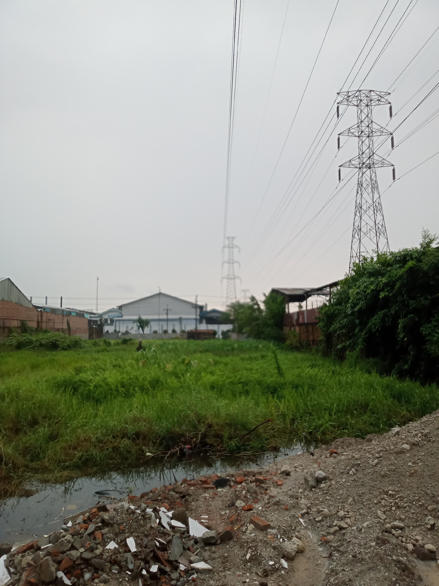 This image shows an industrial area in the city of Sunggal, Indonesia. In the foreground, there is a pile of debris including broken bricks and white plastic pieces, next to a small body of stagnant water with some reflected clouds and greenery. Behind this, a large patch of tall green grass dominates the middle ground. To the left of the grass are several industrial buildings, one with a brick facade and another with a white, corrugated metal roof. In the background, two large electrical towers rise against an overcast sky, with numerous power lines stretching between them and across the scene. Dense green foliage is visible on the right side of the image, bordering a covered, open-sided structure.
