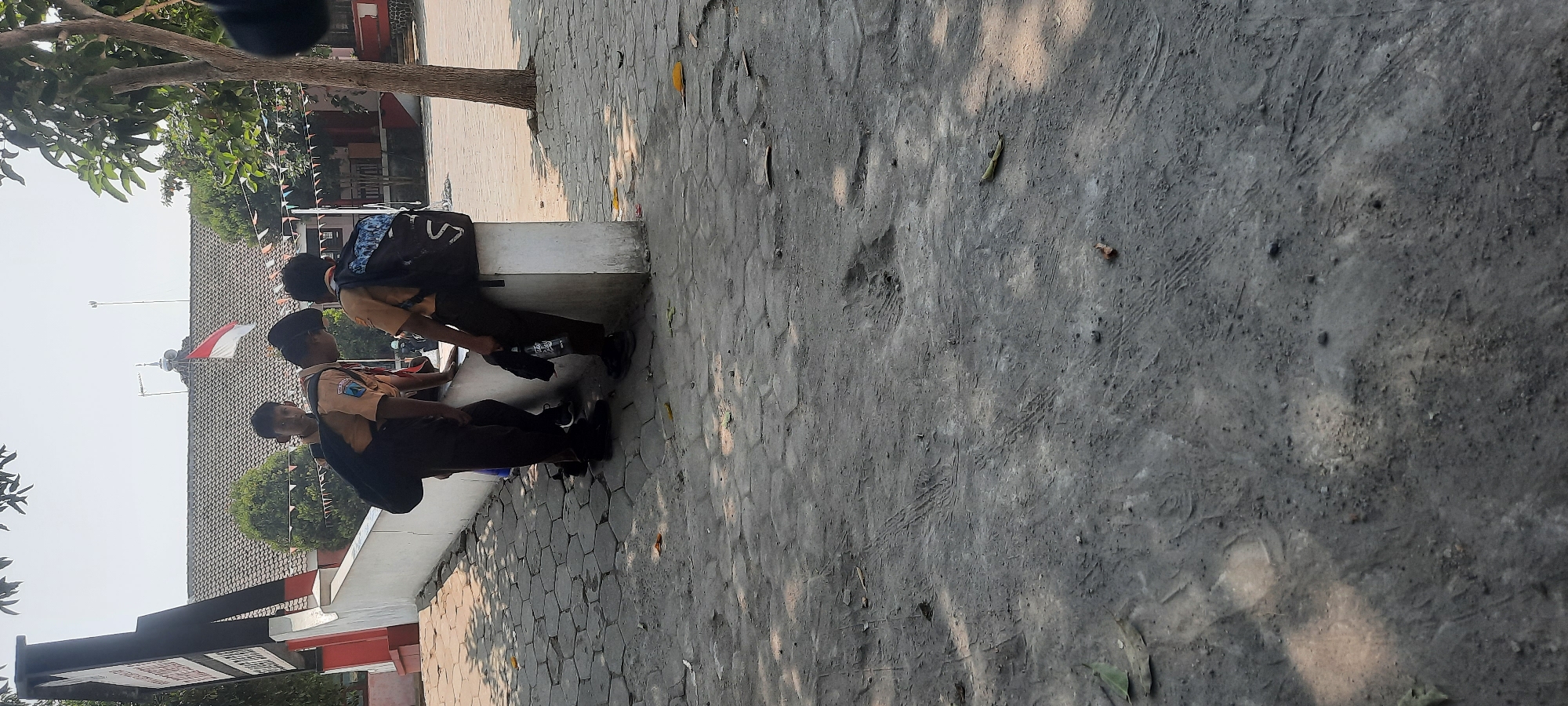 In Tangkil, Indonesia, three young boys in school uniforms and backpacks are gathered near a concrete wall. Two of the boys are standing, and one is sitting on the wall. The boy on the right is holding a black water bottle. In the background, a school building with a red and white Indonesian flag flying and banners decorating its exterior is visible. The ground in front of the boys is paved with hexagonal stones, and the foreground is a rougher, unpaved surface with tire tracks and scattered leaves. The scene is lit by bright sunlight, casting shadows on the ground.