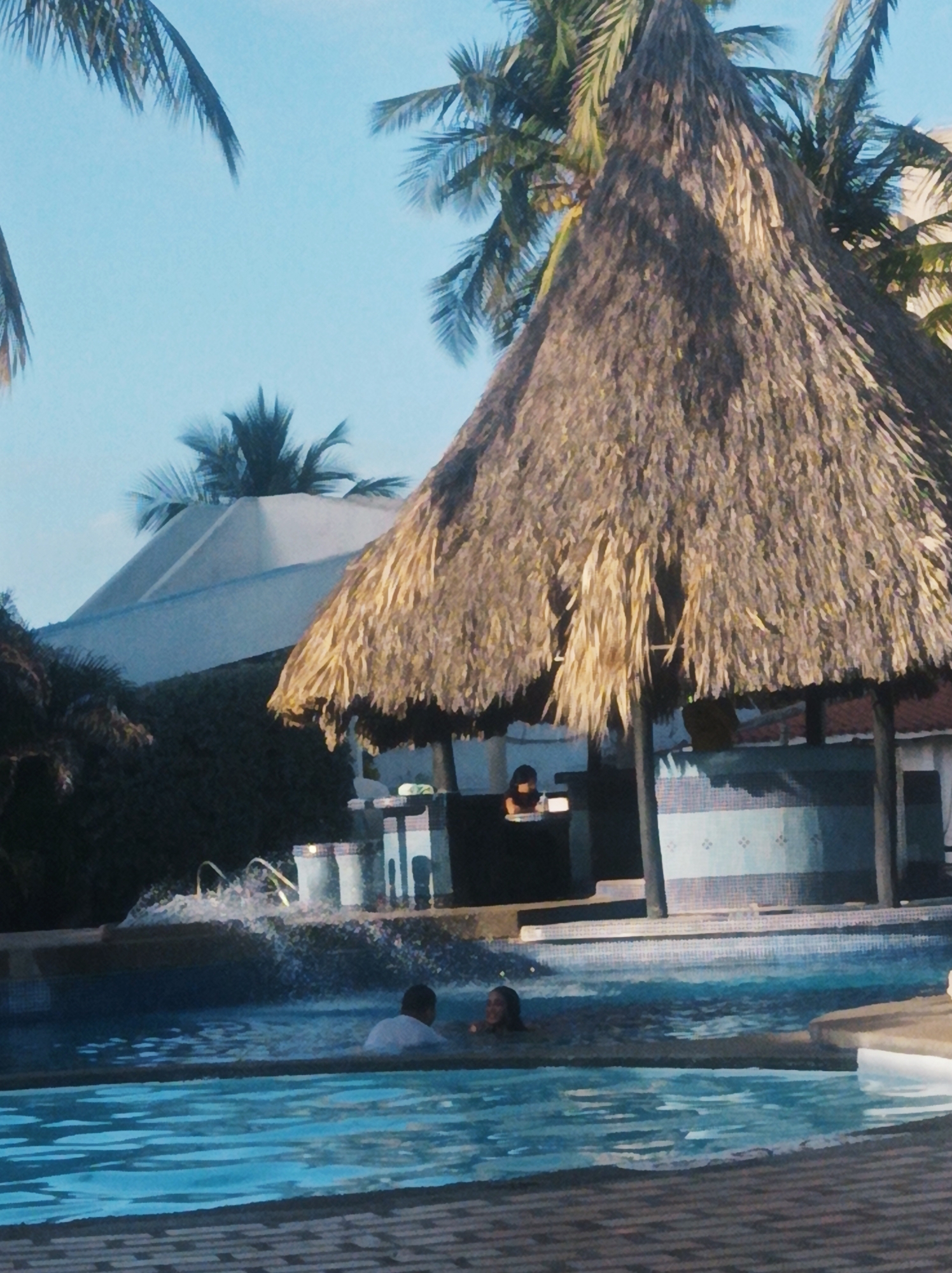 The image depicts a swimming pool scene, likely in a tropical setting. Two individuals are in the pool; one is wearing a white shirt, and the other appears to be a woman. The pool has a visible water feature causing splashing.
Behind the pool, there's a structure with a thatched roof, possibly a bar or shaded area, with what appears to be a person seated behind the counter. The background includes palm trees, a blue sky, and a white umbrella-like structure.
The location is determined to be in the city of Porlamar, Venezuela.