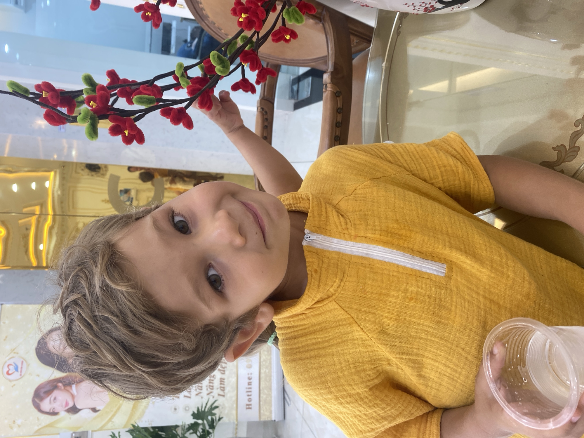 A young child with light brown hair and green eyes smiles into the camera, holding a plastic cup in their right hand. The child is wearing a yellow, textured shirt with a white zipper. The child's left arm is extended, reaching towards a decorative branch with red flowers and green leaves. The branch appears to be made of fuzzy material and is placed in a vase on a table. The table is made of wood with a reflective surface. In the background, there is a display with images of two women and some text. The setting appears to be indoors, possibly a shop or restaurant, given the lighting and the nature of the display. The overall mood is cheerful and playful. Visible text includes "Hotline: 09" and other Vietnamese characters that are partially obscured. The setting suggests it might be daytime due to the bright lighting.