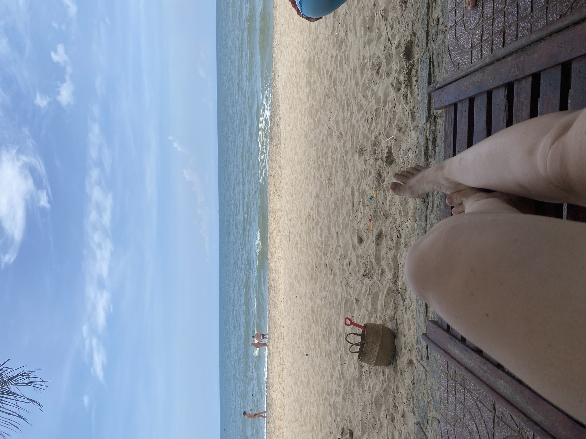 In Phu Thuy Ward, Vietnam, a person lies on a beach chair, their legs and feet in the foreground, facing the sea. The scene depicts a sandy beach with a woven basket and a red shovel in the sand to the left. The ocean extends to the horizon under a partly cloudy sky. In the distance, several people are visible near the water's edge. The overall atmosphere is relaxed, suggesting a vacation or leisurely day at the beach.