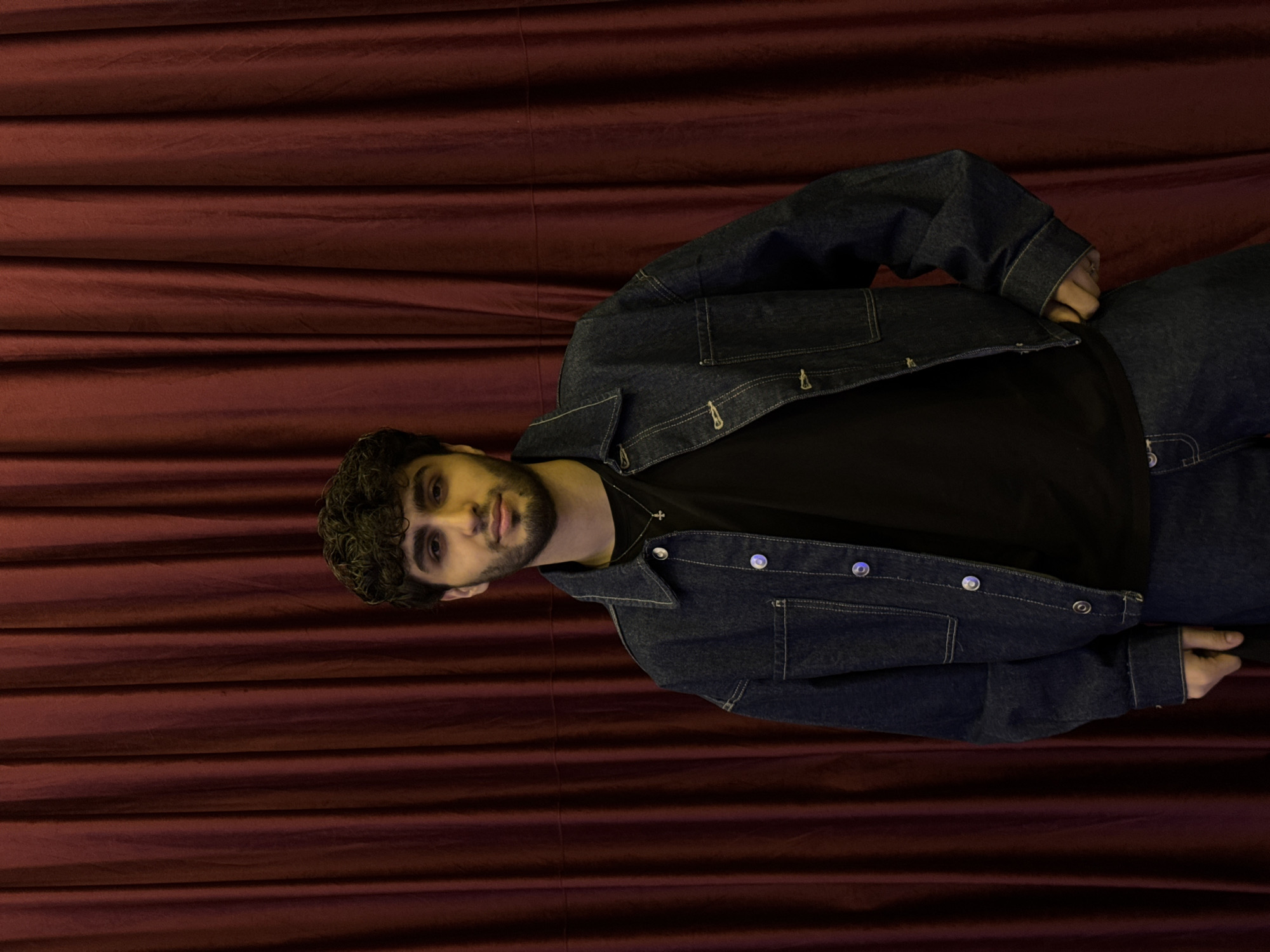 A man is lying down against a maroon, horizontally draped curtain backdrop. He has dark hair and a beard, and he's wearing a black t-shirt under a dark blue denim jacket and pants. He appears to be posing. The location is undetermined, and there is no evidence to suggest this picture was taken in Yerevan, Armenia.