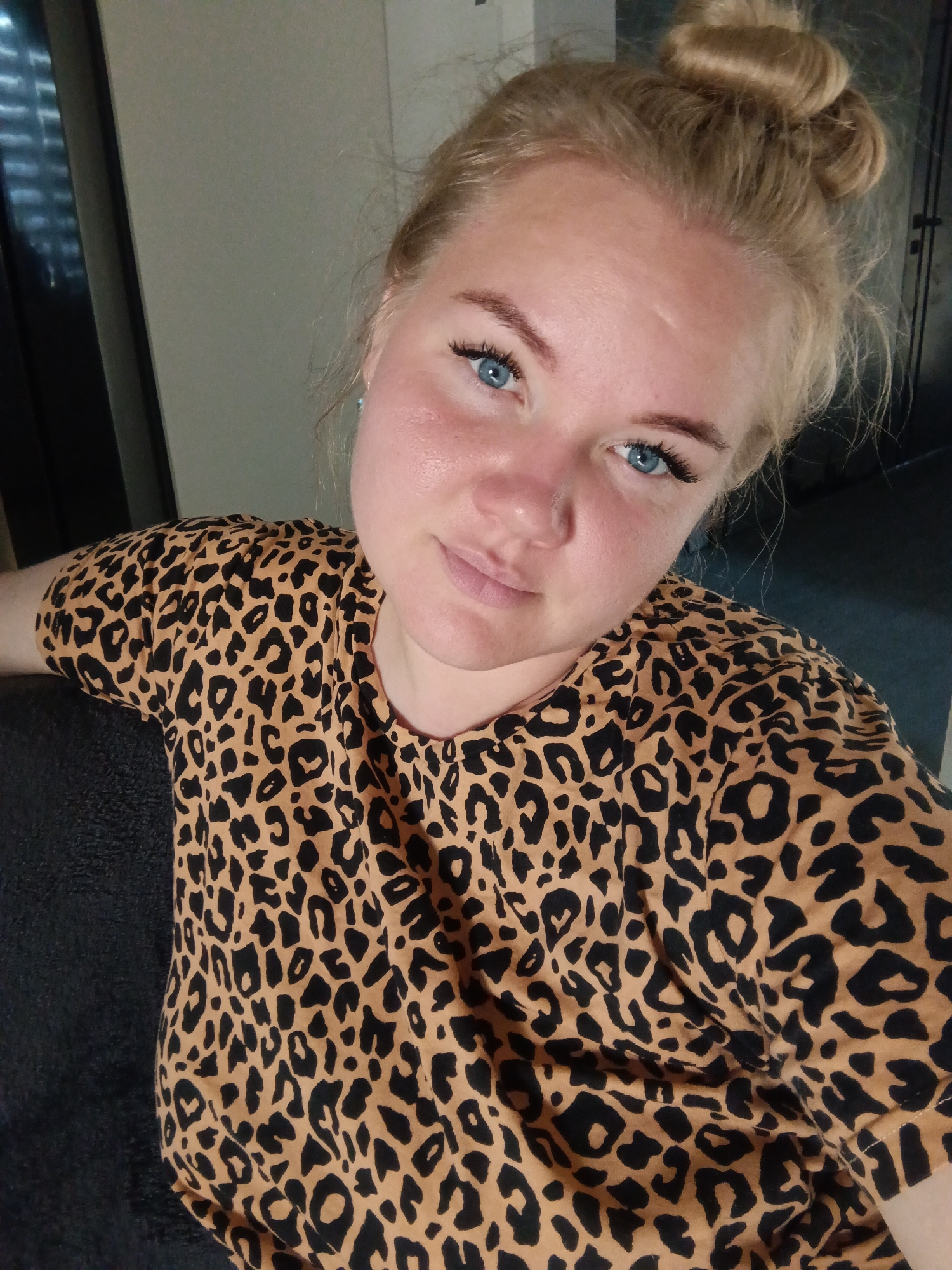A blonde woman with blue eyes and her hair in a bun is shown from the chest up, looking towards the camera. She is wearing a leopard print t-shirt and is sitting down, with her left arm resting on a dark-colored surface. The background is out of focus, showing a wall and what appears to be a window with blinds. There is no specific indication of the location being in Фоки, Russia.