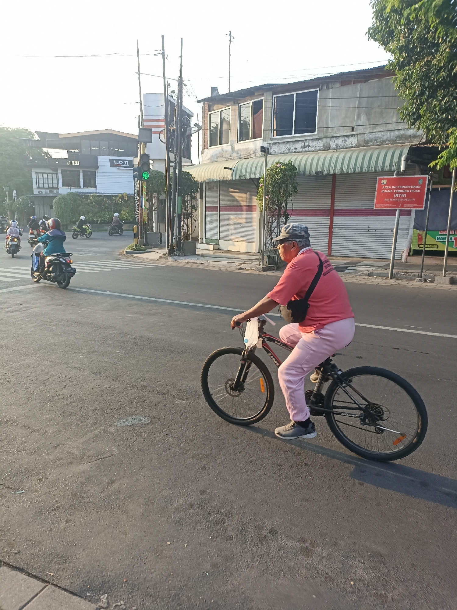 In Mojopanggung, Indonesia, an elderly man in a camouflage hat, pink t-shirt, and pink pants rides a black bicycle. He is wearing gray athletic shoes and has a black bag slung across his chest. The bicycle has a black frame and silver handlebars. The man is in the foreground, on the right side of the image, and is moving from left to right. The background shows a street with several motorbikes and their riders, suggesting a busy intersection. Traffic lights are visible, with one showing green. Buildings line the street, some with awnings. The sun is low in the sky, casting long shadows.