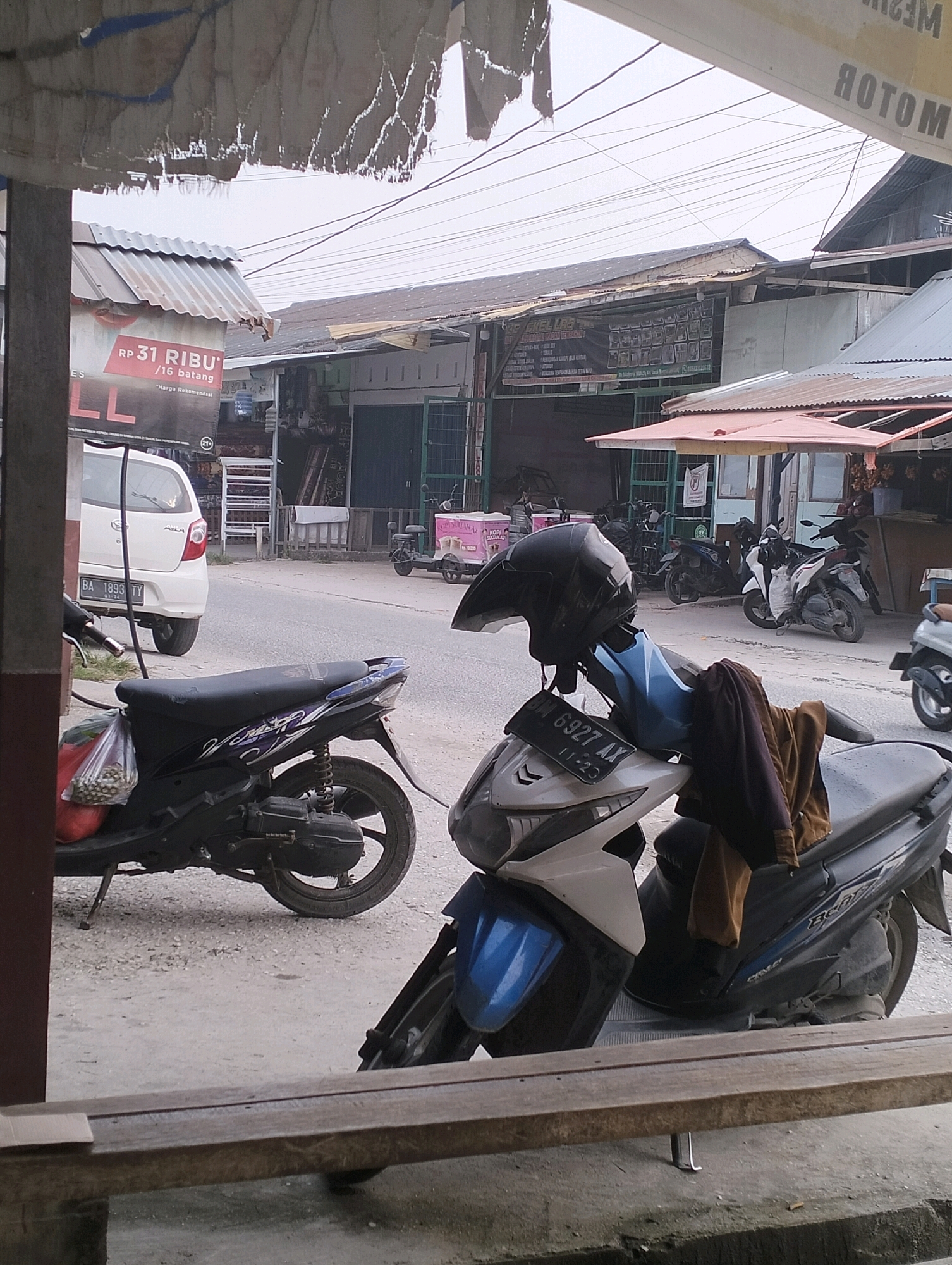 The image depicts a street scene in Tarai Bangun, Indonesia, with multiple parked motorcycles and a white car. In the foreground, a dark blue and black scooter is parked on the left. On the right, a white and blue scooter is parked with a black helmet and brown clothing draped over its seat. A white car with the license plate "BA 1893 TY" is parked on the left side of the street. In the background, there are several shops and buildings lining the street. The sky is overcast, and the overall lighting suggests it is daytime. There are no people visible in the frame.