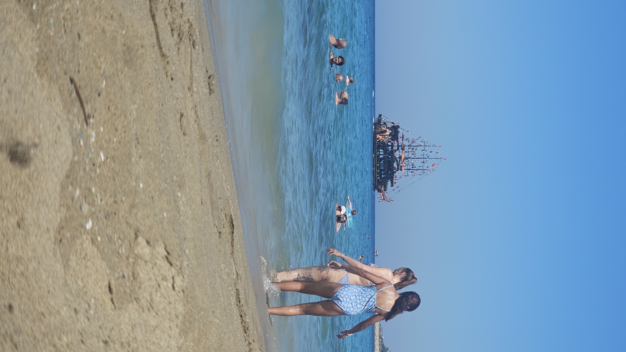The image depicts a sunny day at a beach in Alanya, Turkey, with several people enjoying the water. In the foreground, the sandy beach meets the turquoise water of the Mediterranean Sea. Two young females, one in a blue floral swimsuit and the other in a bikini, are standing at the water's edge, facing away from the camera. In the middle ground, a group of individuals are bathing and swimming in the sea. Further in the distance, a pirate-themed ship is sailing on the clear blue water. The sky is a vibrant, cloudless blue. The overall scene is one of leisure and enjoyment on a warm day at the coast.
