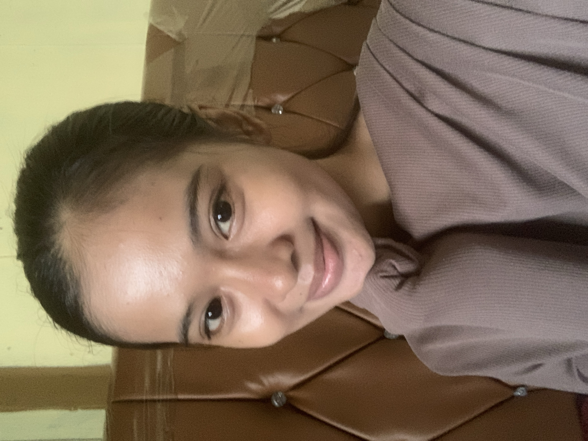 A young woman with dark hair tied back is pictured in a close-up, selfie-style portrait. She has medium-toned skin and is looking towards the camera with a slight smile. She is wearing a muted purple, ribbed knit top. The background consists of a brown upholstered headboard with decorative studs and a light yellow wall. The image appears to have been taken indoors, likely in a bedroom.