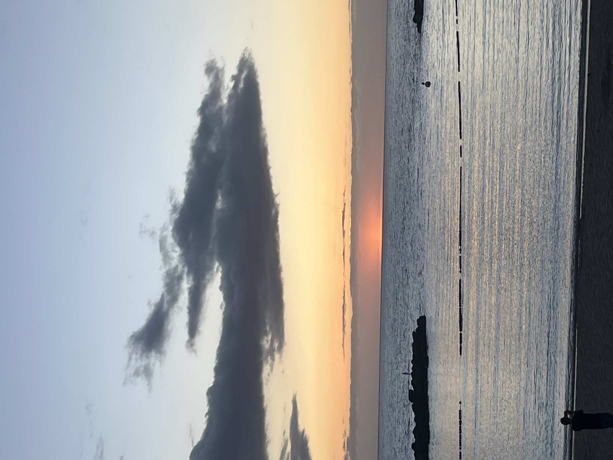 The image shows a sunset scene at the coast. A large, dark cloud formation is visible on the left side of the image, contrasting with the lighter, orange-hued sky. The sun is setting on the horizon, creating a visible line where the sky transitions to the ocean. The water appears calm with ripples, and the light is reflecting in the water, creating a shimmering effect. Along the shoreline, a breakwater with rocks is visible. On the lower right corner, a person is visible on the beach, seemingly observing or photographing the sunset. The location is likely near the coast of Mogán, Spain, given the presence of the ocean, breakwater, and the characteristic sunset colors.