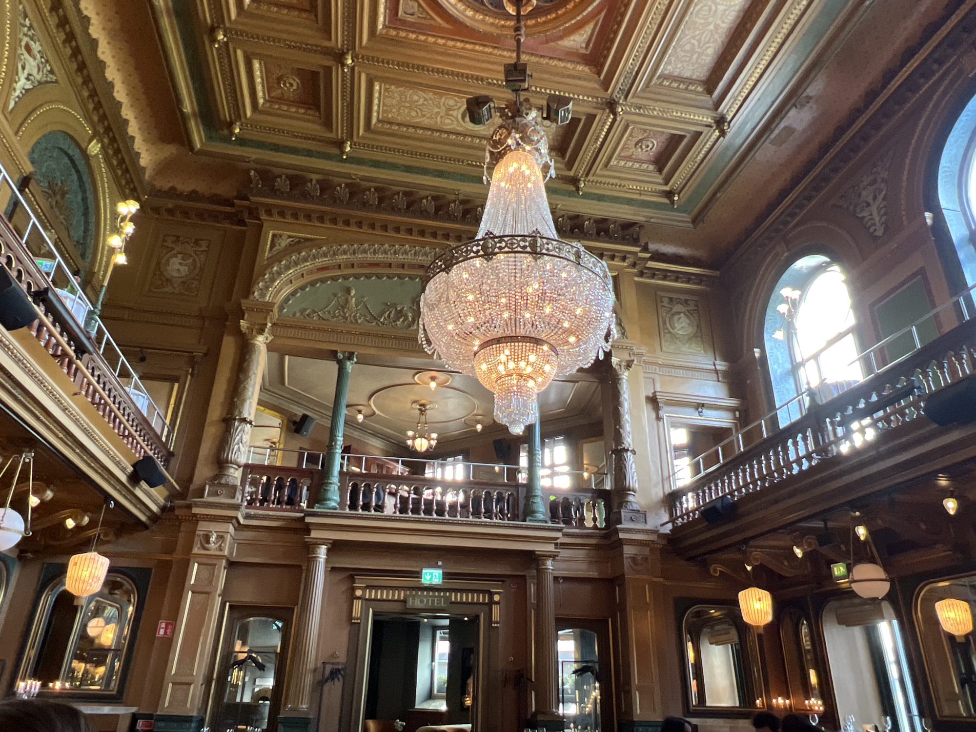 This is an interior shot of the Grand Hôtel in Stockholm, Sweden. The image captures the opulent ceiling and a grand crystal chandelier. The ceiling is ornately decorated with gold accents and intricate moldings, with a large circular central feature directly above the chandelier. The chandelier itself is multi-tiered and made of numerous glass crystals, emitting a warm light. Below the ceiling, a mezzanine level with wooden railings and columns is visible, suggesting a grand hall or ballroom. Windows with sunlight streaming through are visible in the upper right. The lower part of the image shows more of the hotel's interior, including wooden paneling, decorative doorways, and more light fixtures. The overall impression is one of luxury and historic grandeur.