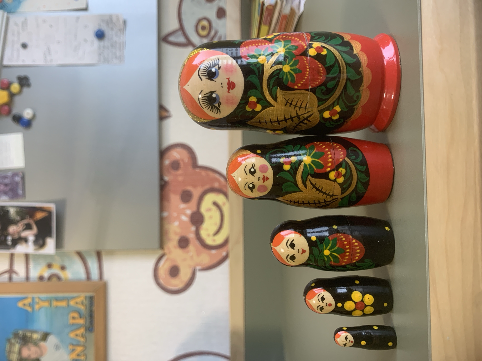 The image shows a set of five Russian nesting dolls (matryoshka dolls) arranged in a row from largest to smallest. The dolls are decorated with traditional Russian folk art patterns, including floral motifs and red accents. The backdrop includes a section of a refrigerator door with various magnets and papers attached, a framed picture or poster, and a wall with a stylized image of a bear. It is not possible to determine if this picture was taken in the city of Izhevsk, Russia.