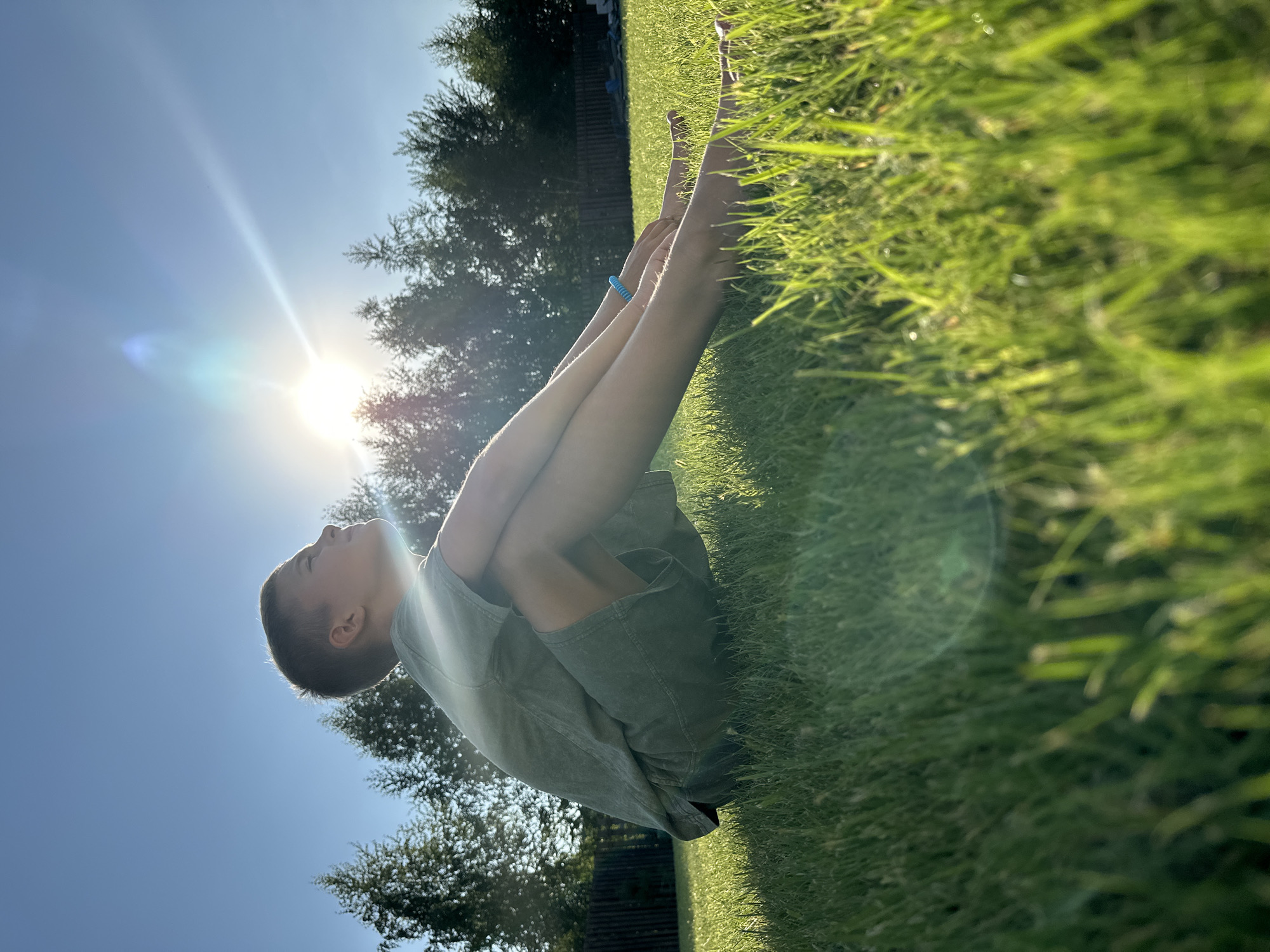 A young boy with short brown hair sits cross-legged in a grassy field, facing the sun. The sun is high in the bright blue sky, casting a lens flare across the image. The boy is wearing a green t-shirt and shorts. In the background, a line of trees forms a dark silhouette against the sky, and a wooden fence is visible. The grass in the foreground is in sharp focus, with some blades catching the sunlight. The overall scene suggests a warm, sunny day, possibly in a suburban or rural area, like the outskirts of the city of Samarsky, Russia, given the presence of trees and a fence.