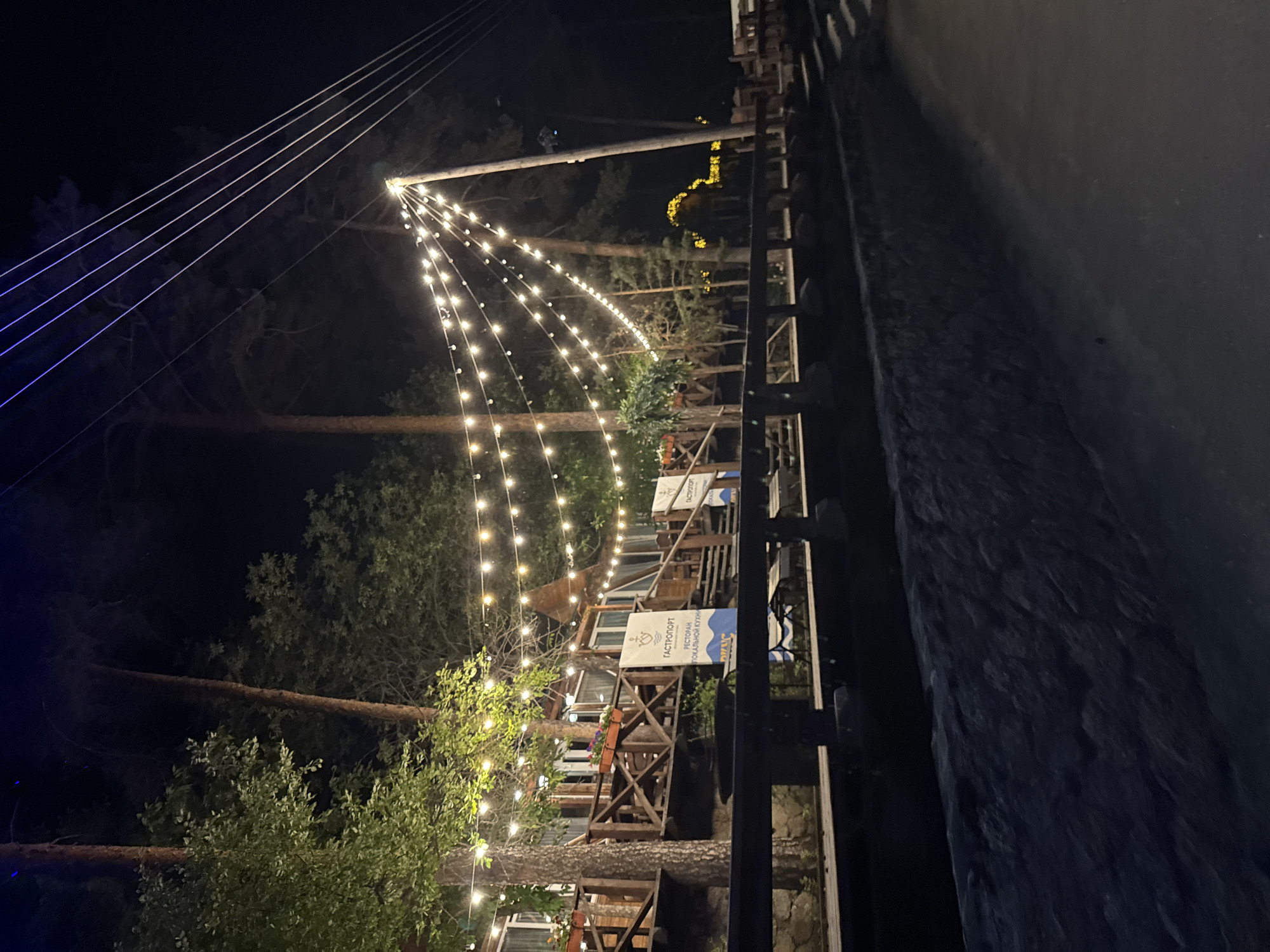 The image depicts an outdoor scene at night, possibly in the city of Zolotoi Plyazh, Russia. A wooden building complex, likely a restaurant or cafe, is visible with several levels and balconies. String lights are strung across the scene, illuminating the area with a warm glow. The building is surrounded by tall trees, and in the foreground, there is a dark, paved area and a stone wall. Two blue and white banners with Cyrillic text are displayed on the building. The lighting suggests an evening ambiance, with the lights creating reflections and highlights on the surfaces. No people are clearly visible in the image.