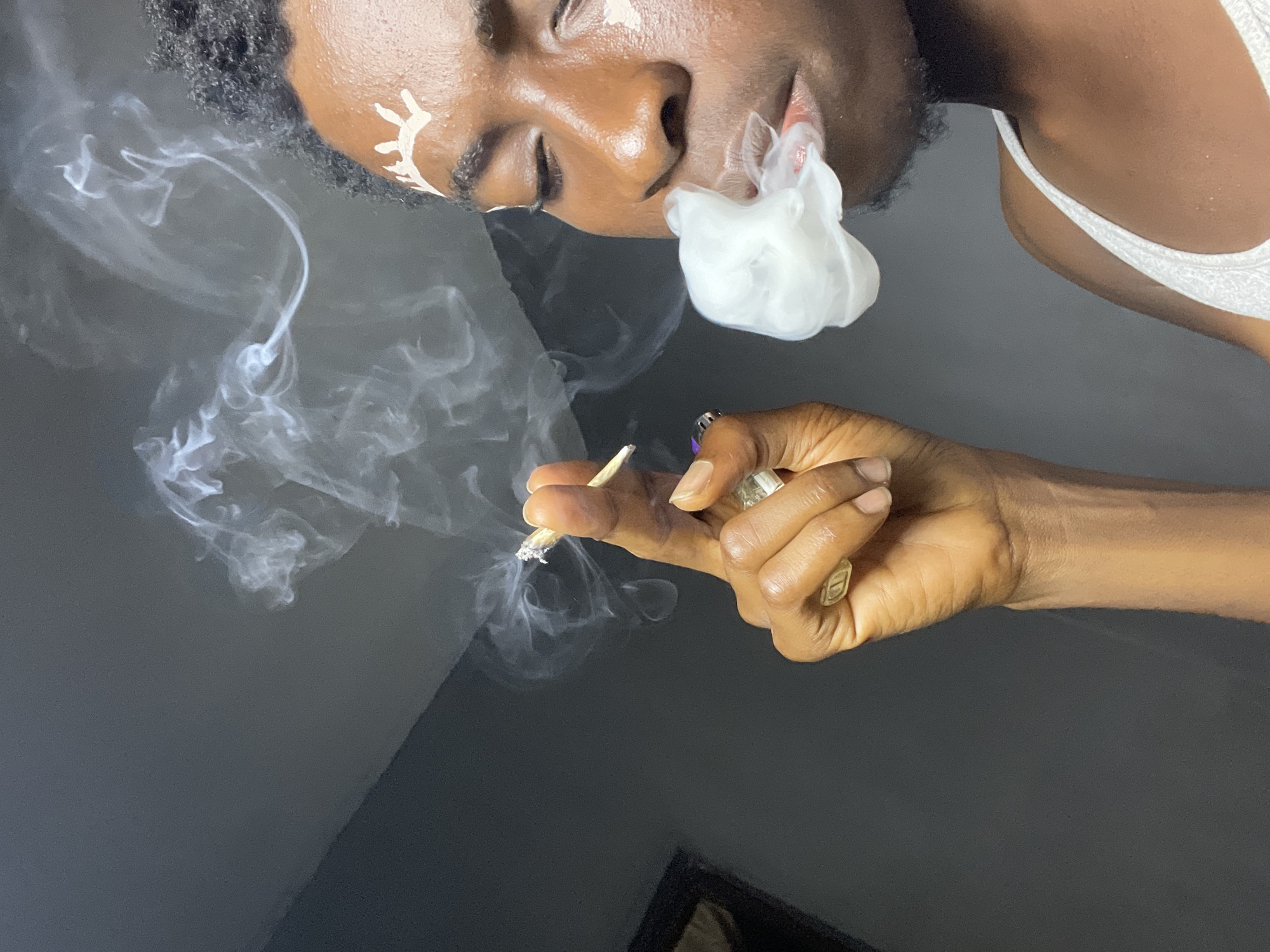 A person is shown in profile, exhaling a large cloud of white smoke from their mouth. Their eyes are closed, and their head is tilted slightly upwards. The person has dark skin and is wearing white face paint in a crown-like design on their forehead. They are holding a lit cigarette between the thumb and index finger of their right hand, which is raised towards their face. The cigarette is small and appears to be burning. The background is a dark, plain wall, with a diagonal line of shadow separating the upper and lower portions. The image has a shallow depth of field, with the person's face and the smoke being in sharp focus, while the background is slightly blurred. There are no clear indications of the city of Nkpor, Nigeria, in the image.