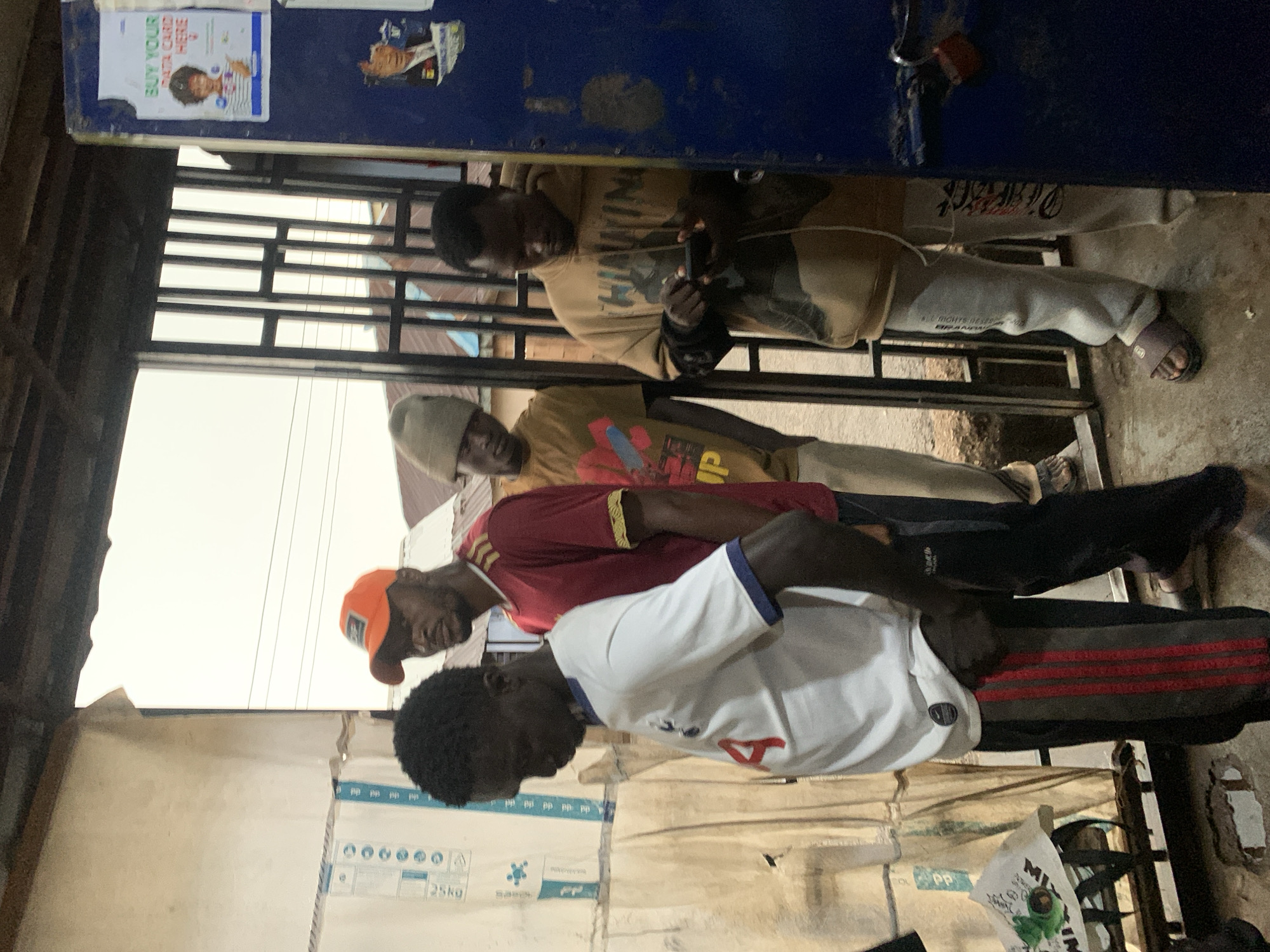The image depicts a street scene in Jos, Nigeria. Four young Black men are the primary subjects, standing near an open doorway or entryway. The man in the foreground, on the left, is wearing a white t-shirt with a blue collar and trim, and black pants with red and grey stripes. He is looking down, and his arms are bent. Behind him, to the right, is a man in an orange baseball cap and a red t-shirt with yellow trim. He is looking towards the left of the frame. Further back and to the left is a man wearing a light brown beanie and a tan t-shirt with a graphic design. He is looking towards the left. The man furthest to the right is wearing a tan hooded sweatshirt with text and graphics on it, and light grey sweatpants. He is holding a mobile phone and appears to be typing or looking at it. He is also wearing sandals. The environment appears to be an outdoor market or commercial area, with corrugated metal roofing visible overhead and what looks like a blue painted stall or structure to the far right. Posters are affixed to the blue structure, one of which advertises "BUY YOUR DATA CARD HERE." The ground is paved with concrete. The overall lighting suggests it is daytime, possibly overcast.