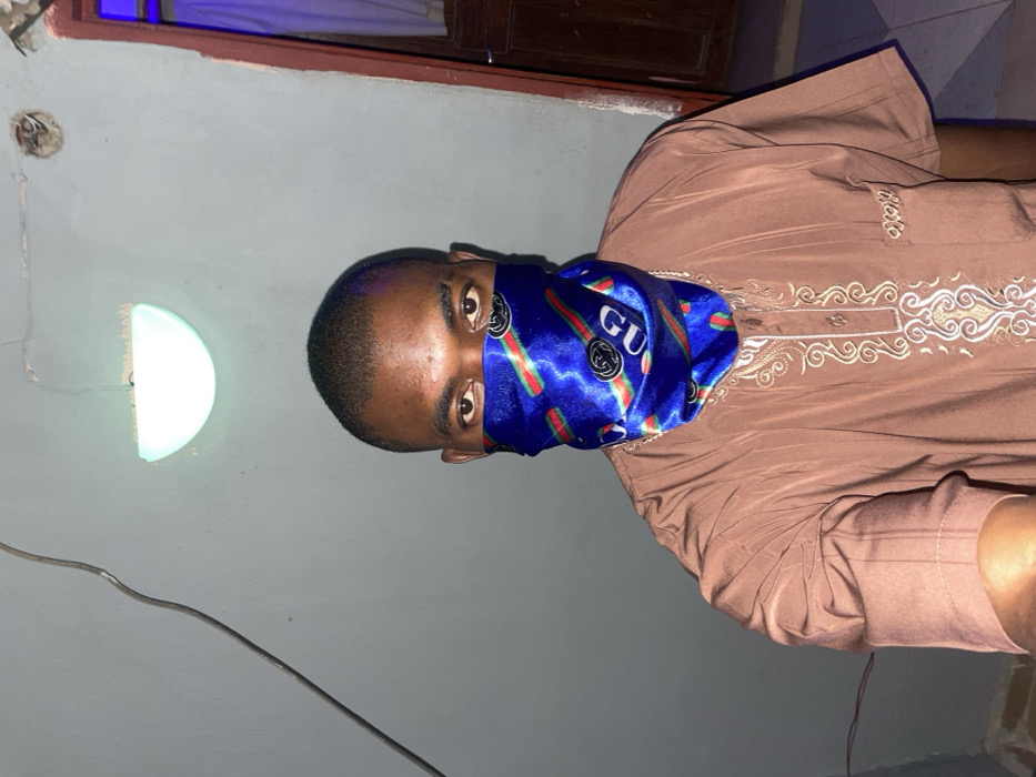 A person wearing a brown embroidered shirt and a blue patterned face covering stands in a room. The covering obscures their nose and mouth. A half-moon-shaped light fixture is on the wall behind them, along with exposed wiring and an electrical outlet. A brown door is visible in the upper background.
