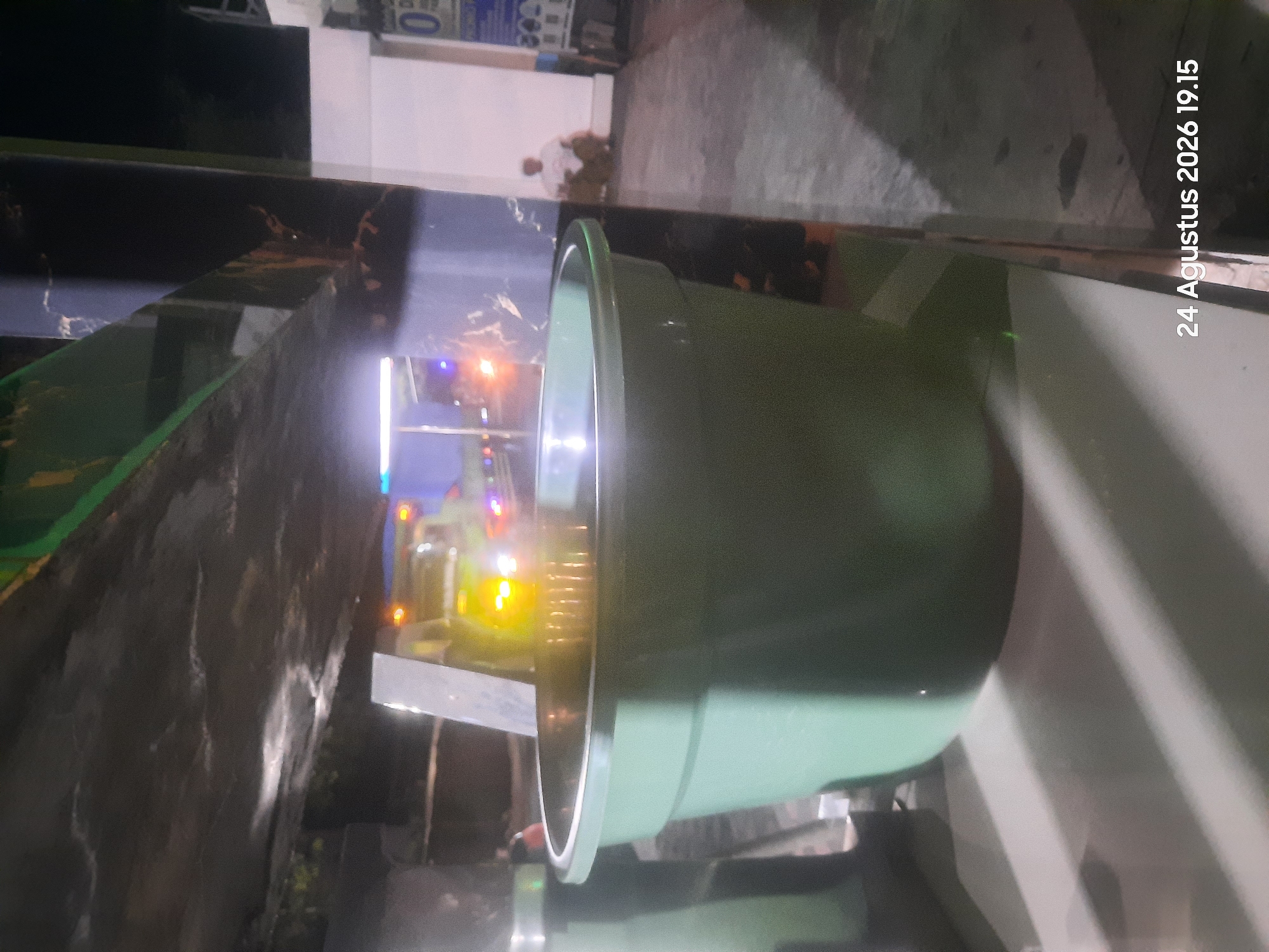 The image captures a nighttime scene in Sukokerto, Indonesia. In the foreground, a large, light green plastic tub is centrally positioned. The tub appears to be filled with a clear liquid, possibly water, reflecting the surrounding lights. In the background, out of focus, is a yellow vehicle with flashing lights, likely an emergency vehicle, parked on a paved area. To the right of the vehicle, a person is seated on the ground in front of a white building. The ground surface is a mix of concrete and asphalt. Shadows from overhead structures are cast across the scene, indicating artificial lighting. A timestamp at the bottom right corner reads "24 Agustus 2026 19.15".