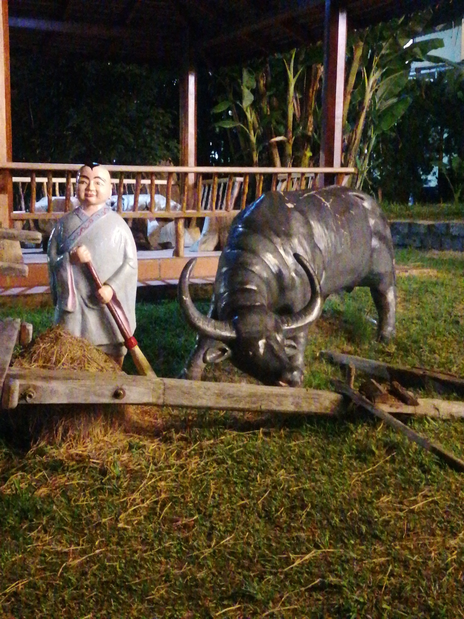 The image depicts a garden scene at night, likely in Da Lat, Vietnam, illuminated by artificial light. In the foreground, a dark, life-sized statue of a water buffalo with prominent horns is positioned to the right. To the left of the buffalo, a statue of a person, possibly a monk or a young boy, is dressed in traditional grey robes and holds a broom with a red handle. The statues are situated in a grassy area with scattered dry straw or hay, suggestive of a rural or agricultural theme. Behind the figures, a wooden structure with a thatched roof and a railing is visible, and beyond that, dark foliage and trees form the background. The overall atmosphere is serene and somewhat mysterious due to the nighttime lighting.