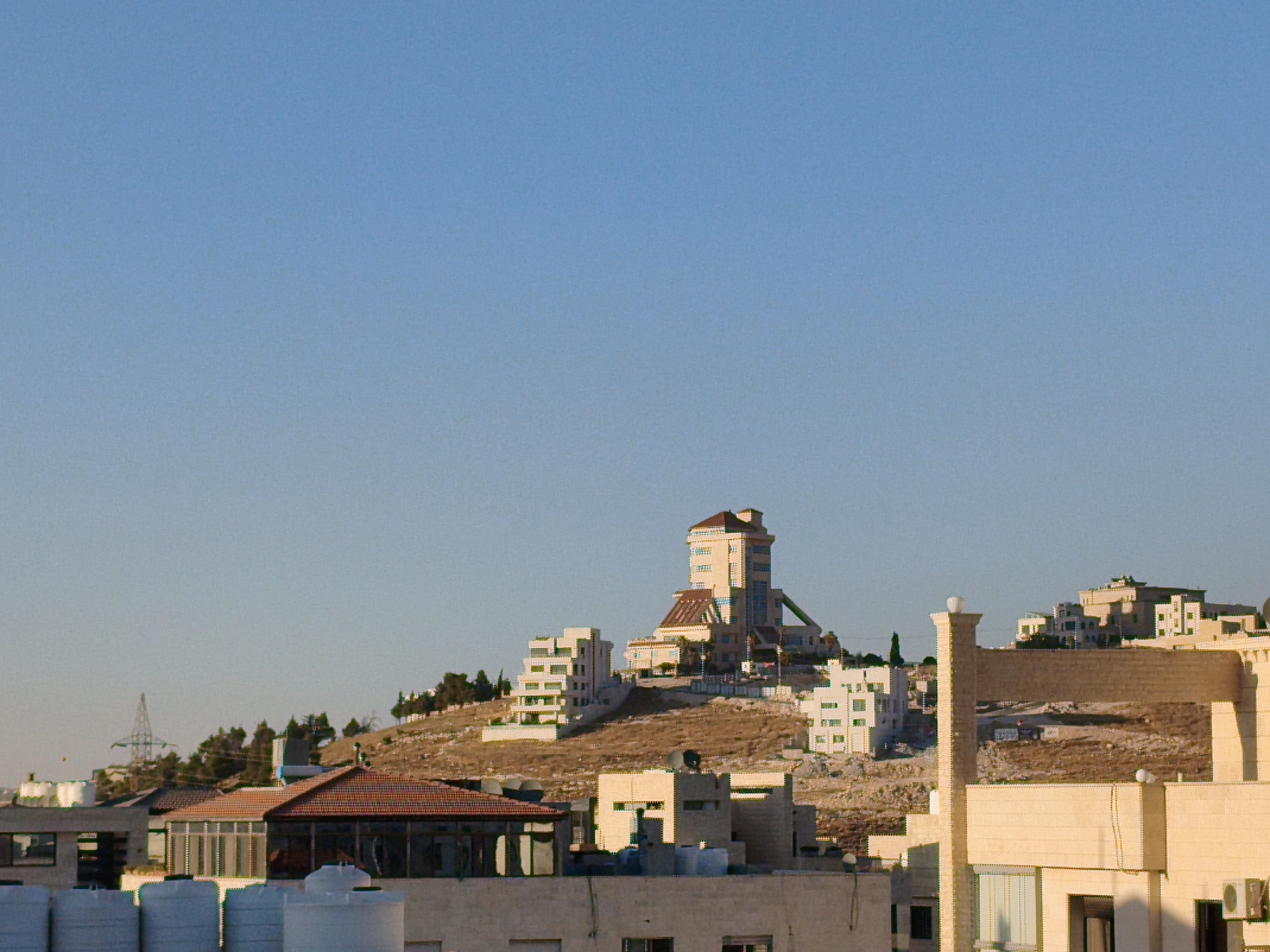 This image depicts a cityscape of Amman, Jordan, with a focus on buildings constructed on a hillside under a clear blue sky. The central feature is a tall, distinctive building with a stepped facade and a pointed roof, situated prominently on the elevated terrain. To its left, a modern white building with a terraced design cascades down the slope. Other residential and commercial structures, varying in size and architectural style, are scattered across the hillside and in the foreground. Some buildings feature red-tiled roofs, while others are painted in neutral tones. In the distance, a transmission tower can be seen against the horizon. The landscape is characterized by arid, rocky terrain dotted with sparse vegetation. The scene conveys a sense of urban development integrated with the natural topography of the city.