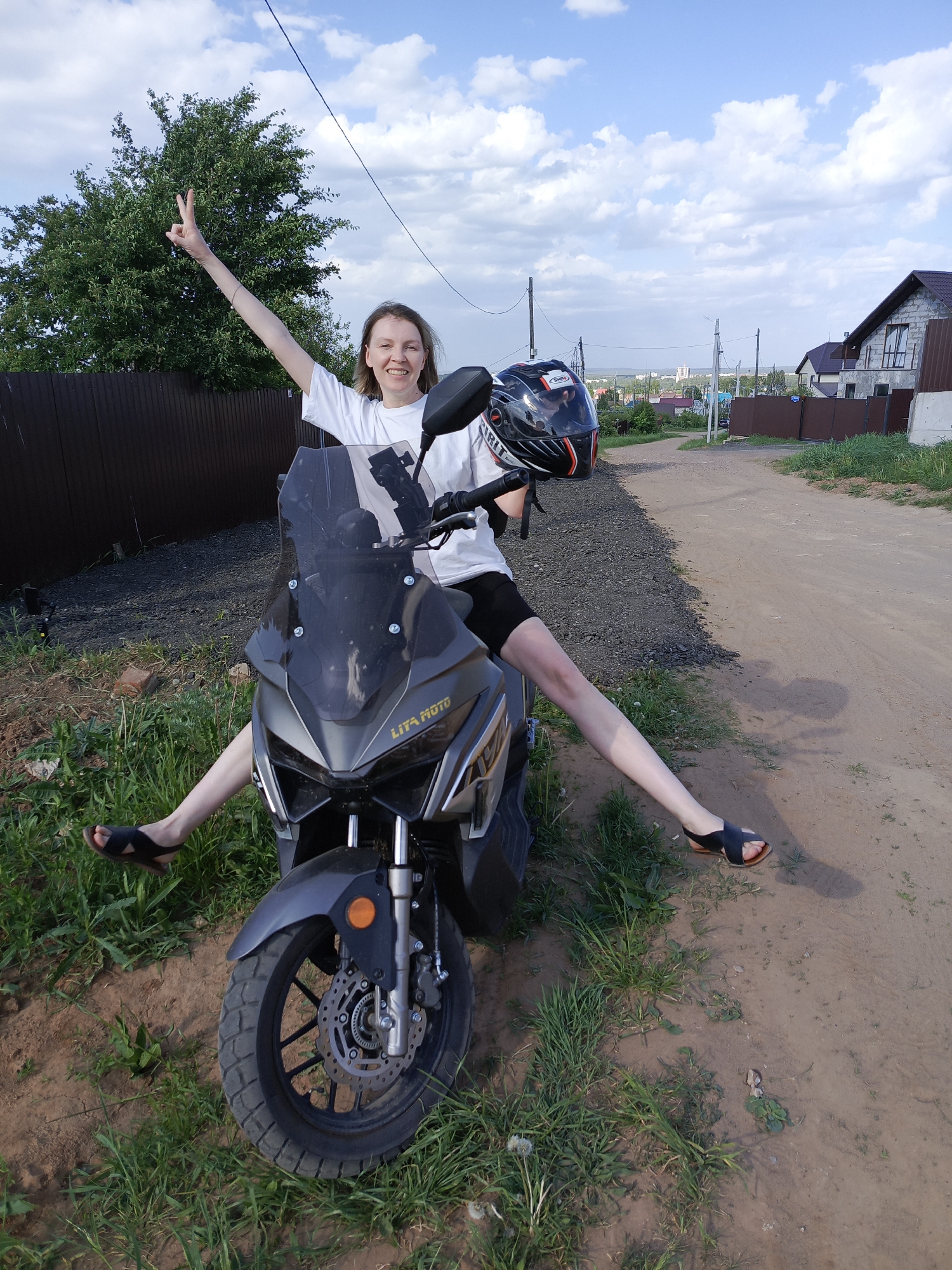 A young woman sits astride a gray Lita Moto scooter on a dirt road in what appears to be a residential area. She is wearing a white t-shirt, black shorts, and sandals. Her left arm is raised, making a peace sign. In her right hand, she holds a black and red helmet. The scooter is positioned on the left side of the road, with grass and vegetation visible around its tires. Behind the woman, a brown fence separates the road from a yard with a tree. Further back, houses and utility poles line the road. The sky is blue with scattered clouds. The location is likely Chaikovsky, Russia based on the scooter brand "Lita Moto".