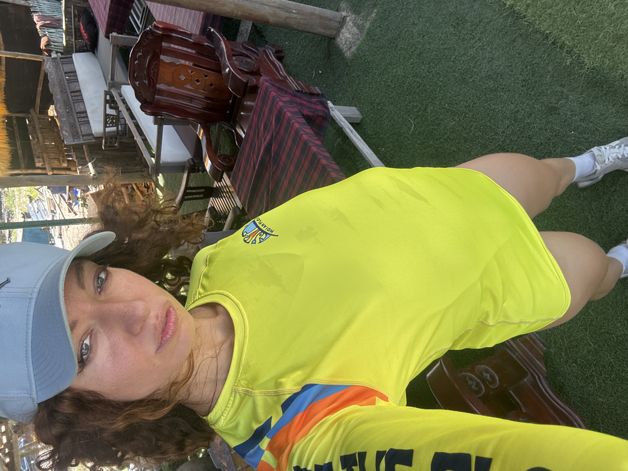 A person wearing a yellow long-sleeve shirt and a gray baseball cap is centered in the frame. The shirt has blue and orange stripes on the sleeve and black lettering that reads "THE" on the lower portion. The person has brown, curly hair and is looking directly at the camera with a neutral expression.
The background is a mix of outdoor and indoor elements. There's a glimpse of a thatched roof structure on the left. In the midground, there are wooden tables and chairs, some with striped red and black table runners. There is grass on the ground.
Given the shirt's inscription "HOIAN FLO", the presence of a structure with a thatched roof, and the outdoor setting, it's reasonable to assume the location is either in or near Hoi An, which is not in Đà Nẵng, but is a neighboring city. The scene appears to be taken in an outdoor seating area of a restaurant or cafe.