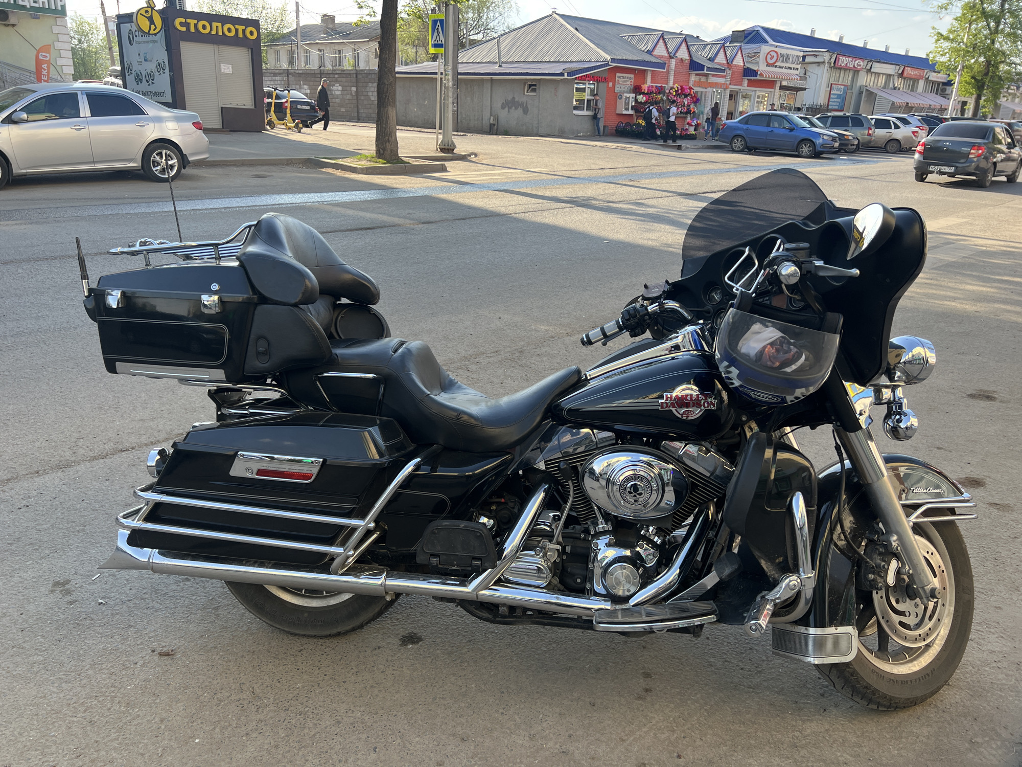 This is an outdoor daytime shot featuring a black Harley-Davidson motorcycle parked on a city street. The motorcycle has a large windshield, chrome engine details, black leather seats, and storage compartments in the back.
In the background, there is a street scene with various buildings and people. On the left, there is a silver car parked near a "СТОЛОТО" kiosk. Further down the street, there is a group of people walking. In the distance, there are shops with red and white facades, and several parked cars. The city in the background is Ufa, Russia.