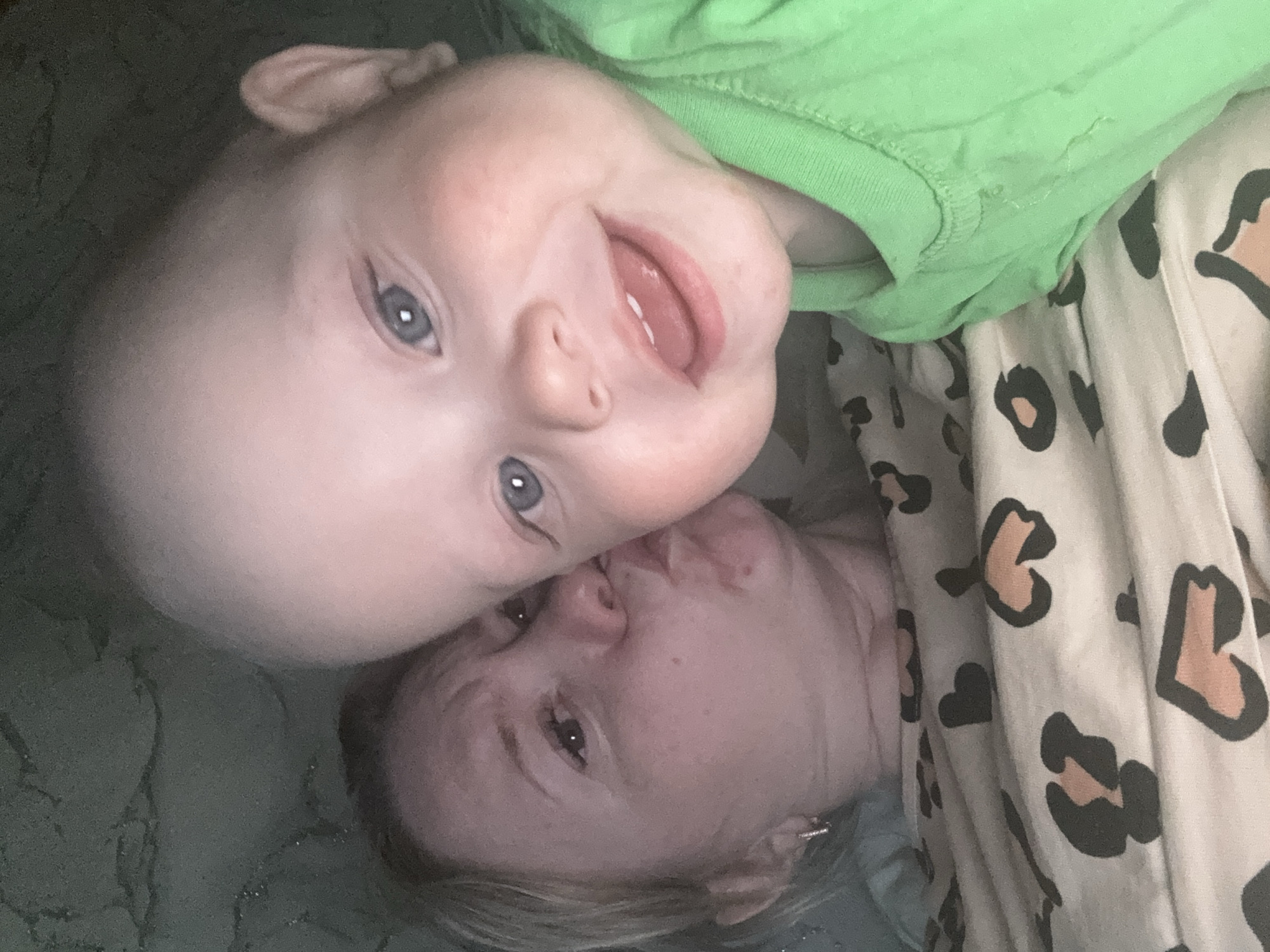 The image shows a baby in a green shirt lying on top of an adult, who is lying on patterned bedding. The baby is smiling and has blue eyes. The adult is looking up towards the camera. The location is not identifiable as Chaikovsky, Russia, based on the visual information in the image. There are no landmarks or other indicators to identify the location.