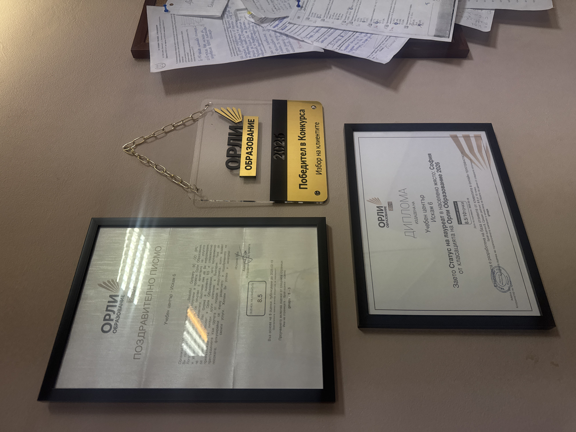This image depicts a section of a wall, where several framed certificates and awards are mounted. These documents appear to be related to an educational institution, with the recurring name "Orli Obrazovanie" indicating a brand or organization involved in education.
The awards and certificates are predominantly in Bulgarian, suggesting the location is in Bulgaria, specifically Sofia as indicated in one of the certificates. The certificates highlight achievements, such as awards for "Winner in the Competition - Choice of Clients," a diploma for "Status of Laureate in Sofia," and a congratulatory letter. They all seem to be associated with a learning center, "Iskam 6." The quality of the printing and framing suggests that these are genuine accomplishments, professionally recognized.
Additional papers are mounted above the awards. The environment is an office.