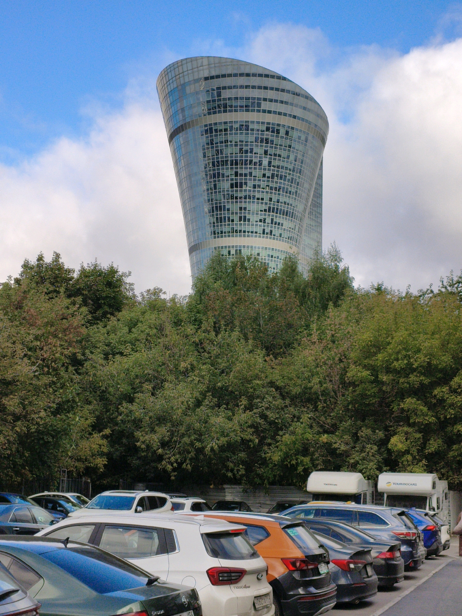 The image depicts the Dominion Tower, a uniquely twisted skyscraper in Moscow, Russia, on an overcast day with patches of blue sky. The building's facade is a grid of reflective windows. In the foreground, a parking lot is filled with various cars, including a white SUV, a dark sedan, an orange car, and a blue hatchback. Two white camper vans with "TOURINGCARS" branding are parked behind the cars. A dense line of green trees partially obscures the view of the parking lot and the base of the skyscraper, suggesting an urban park or a natural buffer zone around the building. The overall scene captures a modern architectural landmark juxtaposed with everyday urban life.