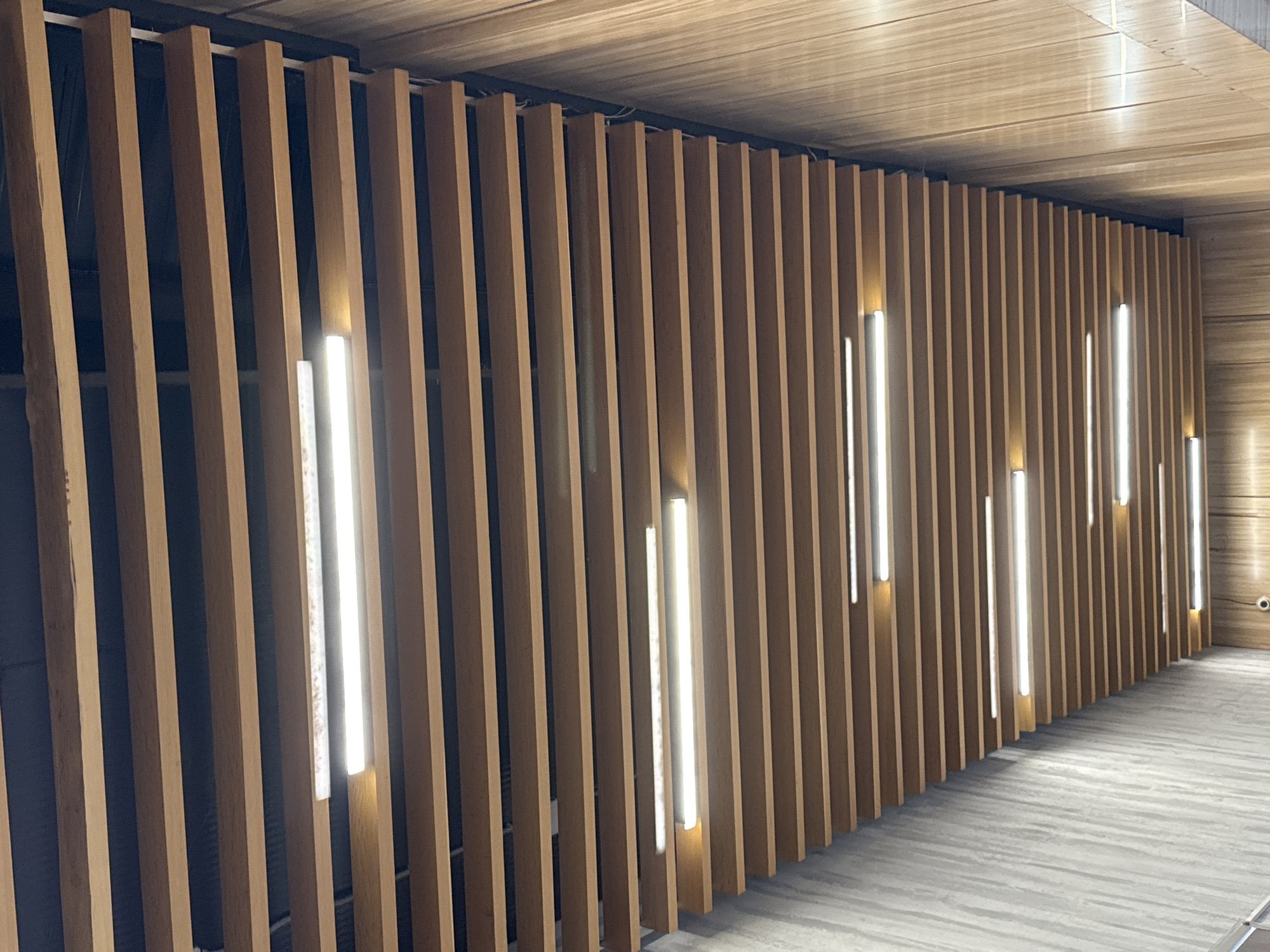 The image shows a long, narrow passageway with a slatted wooden ceiling. Fluorescent lights are embedded between the wooden slats, illuminating the space. The walls of the passageway are made of concrete on the left and wood paneling on the right. There are no people or animals visible in the image. The overall impression is of a modern, indoor architectural space.
Regarding the location in Norilsk, Russia: Norilsk is a city in Krasnoyarsk Krai, Russia, located above the Arctic Circle. It is known for its industrial activity, particularly nickel and palladium mining and production. The architecture in Norilsk often reflects the harsh climate, with many buildings designed for cold weather insulation. The image does not contain any specific visual cues to definitively place it within Norilsk, Russia. However, the style of the passageway with its functional lighting and durable materials could be found in various public or commercial buildings in the city.