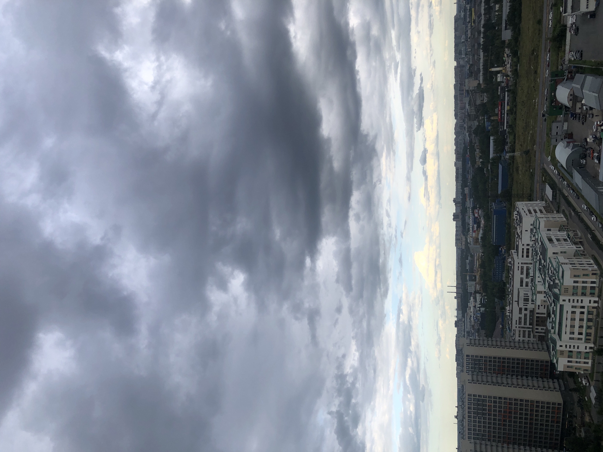 A high-angle, full shot captures an urban cityscape under a heavy, overcast sky. The foreground is dominated by a cluster of modern residential buildings, characterized by their white facades, rectangular windows, and flat roofs. One building on the left is taller and features numerous small, square windows arranged in a grid. To the right, a lower, more sprawling complex of buildings presents a variety of architectural styles.
Beyond the residential buildings, the city stretches into the distance, a dense arrangement of lower-rise structures, industrial buildings, and patches of green space. In the far background, a line of taller buildings forms the urban horizon, punctuated by two prominent industrial chimneys. The sky is filled with dark, heavy clouds, obscuring the sun and creating a somber, diffused light. A few lighter patches in the sky suggest the presence of thinner clouds or breaks in the cloud cover near the horizon. The overall impression is of a sprawling urban environment on a cloudy day.