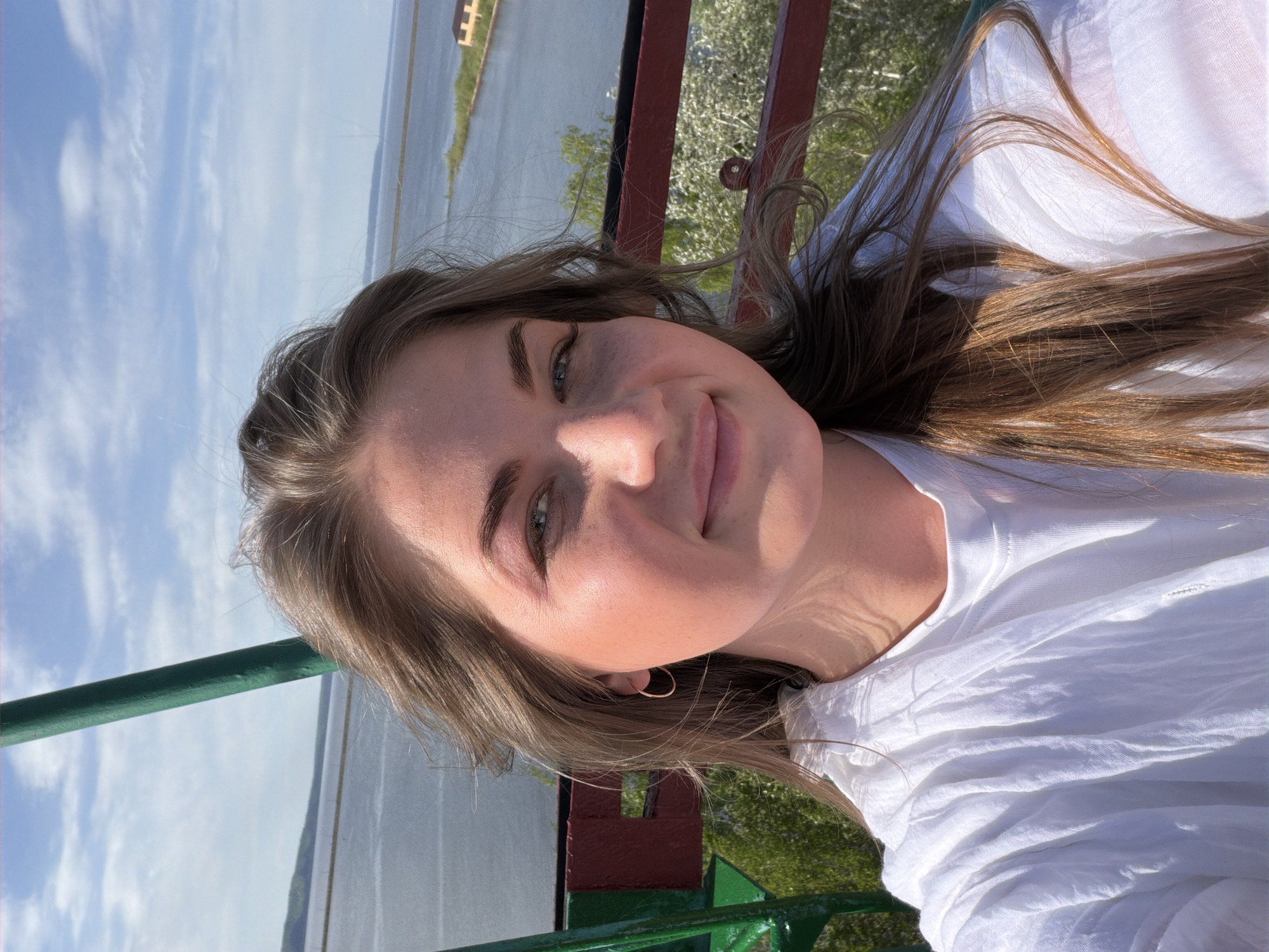 A young woman poses for a selfie outside in daylight. She has long brown hair, fair skin, and is wearing a white shirt. She is smiling slightly. Behind her is a body of water, possibly a river or a lake, and a cloudy sky. There's a railing with green and red paint. The location is potentially Chaikovsky, Russia. The photograph's orientation is somewhat sideways.