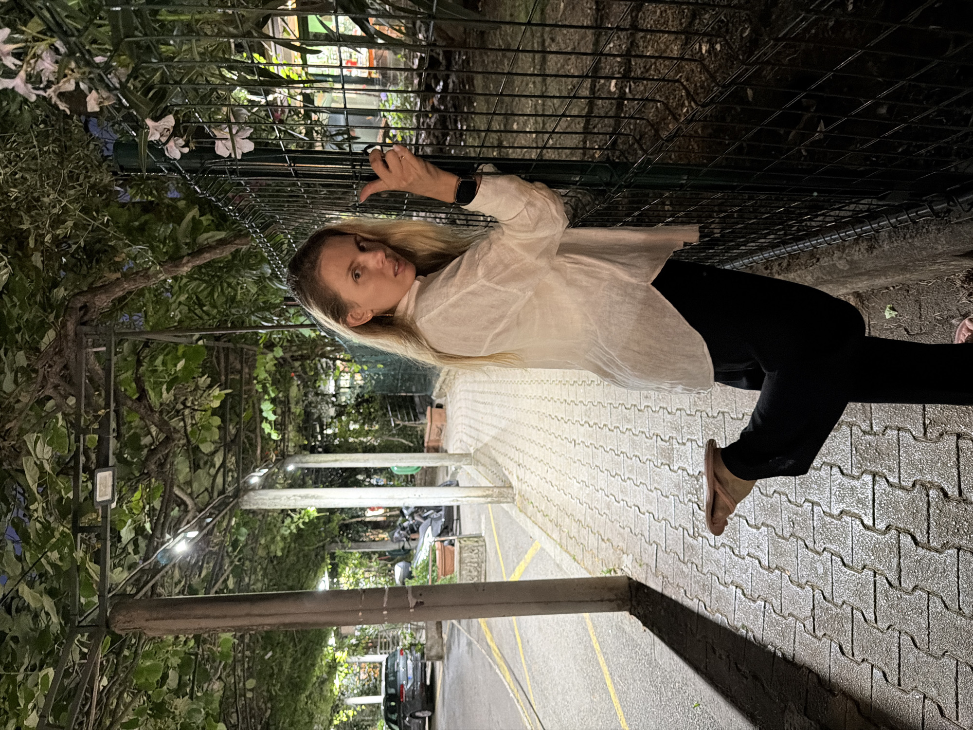 The image captures a woman standing on a paved walkway next to a black metal fence at night, in Budva, Montenegro. She is leaning on the fence with her right hand, while her left leg is bent upwards, with her left foot held in her left hand. She is wearing a white long-sleeved shirt and black pants. The walkway is illuminated by overhead lights, casting shadows on the ground. In the background, trees with green leaves form a canopy, and there is a car and a motorcycle parked on the side of the road. The lighting suggests it is a public area, possibly a park or a promenade.