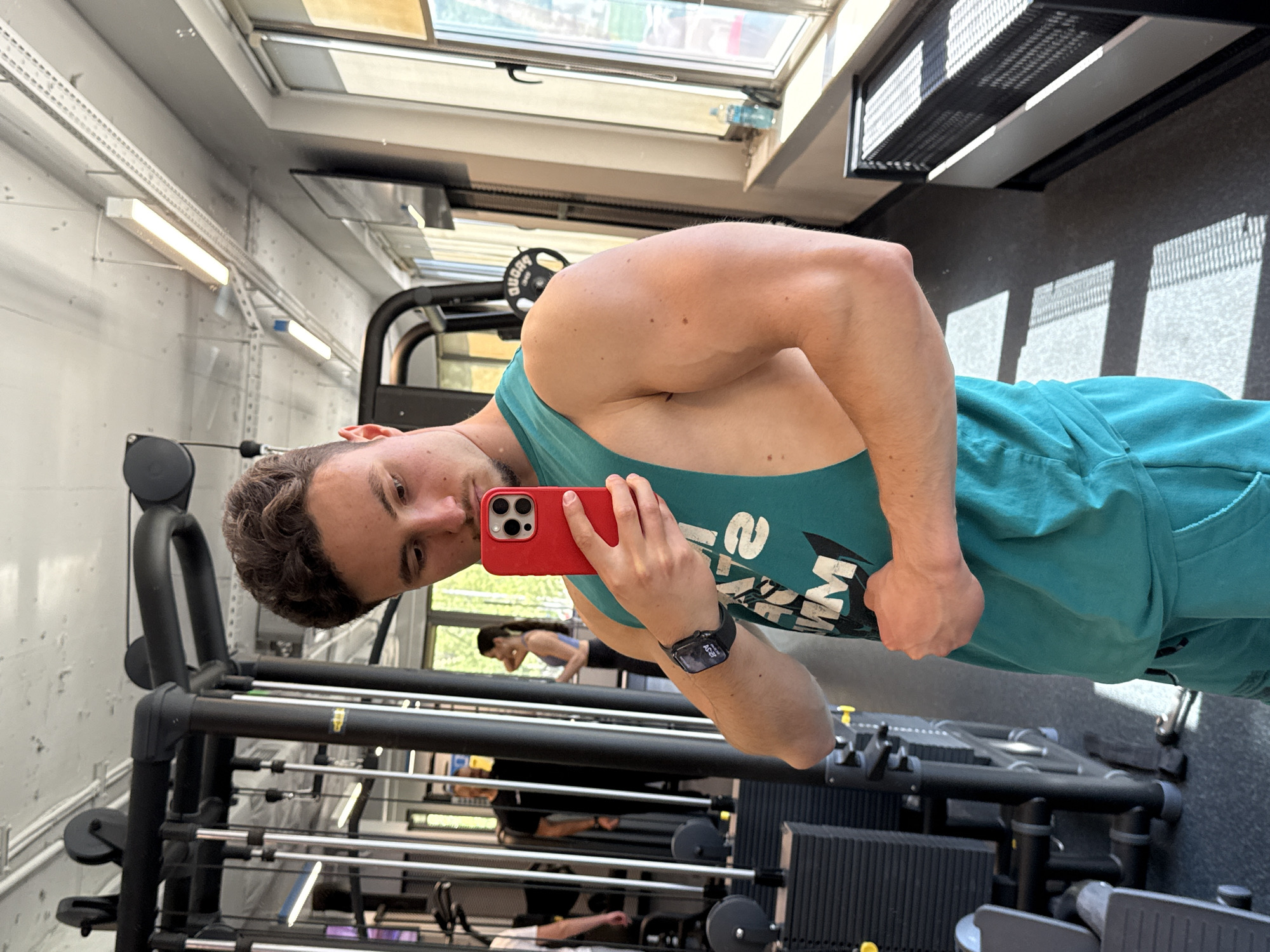 A man is taking a selfie in a gym. He is holding a red smartphone in his right hand. He is wearing a teal-colored tank top and a smartwatch on his left wrist. In the background, exercise equipment is visible, including weight racks and machines. A woman can be seen exercising in the reflection of a mirror behind him. The environment suggests a well-lit, modern gym setting. The location is the city of Sofia, Bulgaria.