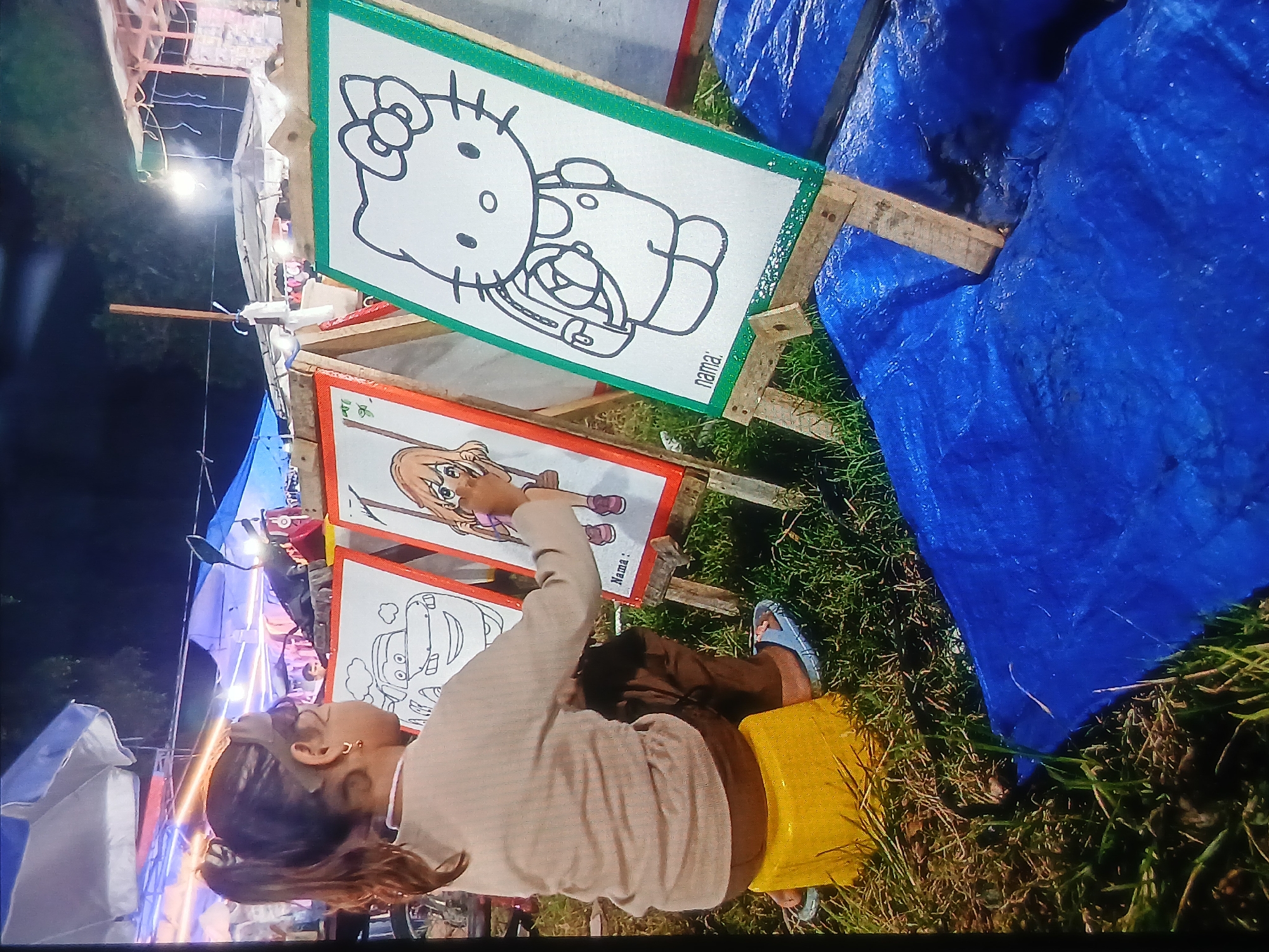This image captures a nighttime scene in the city of Setia Bakti, Indonesia, featuring a young girl engaged in a coloring activity. She is seated on a yellow stool, facing away from the camera, and appears to be coloring on a large sheet of paper displayed on an easel. The easel has a white background with a black outline drawing of a character, possibly a cartoon. The girl is wearing a light-colored long-sleeved shirt and dark pants. Her hair is pulled back into a ponytail. In front of her, other easels are set up, also displaying drawings. One easel shows a black and white outline of a car, and another depicts a character with brown hair. In the foreground, there are blue tarps spread on the ground, and the scene is illuminated by artificial lighting, suggesting an outdoor market or event. The background shows stalls and tents, indicative of a festive or commercial setting at night.
