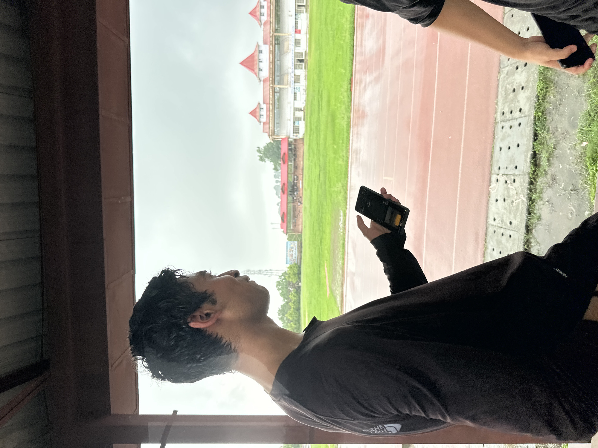 An Asian male in a black long-sleeved shirt stands in profile facing right, holding a black smartphone in his right hand. He is under a covered structure with a corrugated metal roof and a dark brown support beam. His hair is dark and wet, and he is looking down at the phone. In the background, a green field with a red running track is visible. A large white building with red roofs and tiered seating is in the distance. The sky is overcast. Another person's arm and hand holding a black smartphone are visible on the right side of the frame. The ground in the foreground is wet and shows a drainage grate. The overall scene appears to be an outdoor athletic facility under cloudy weather.