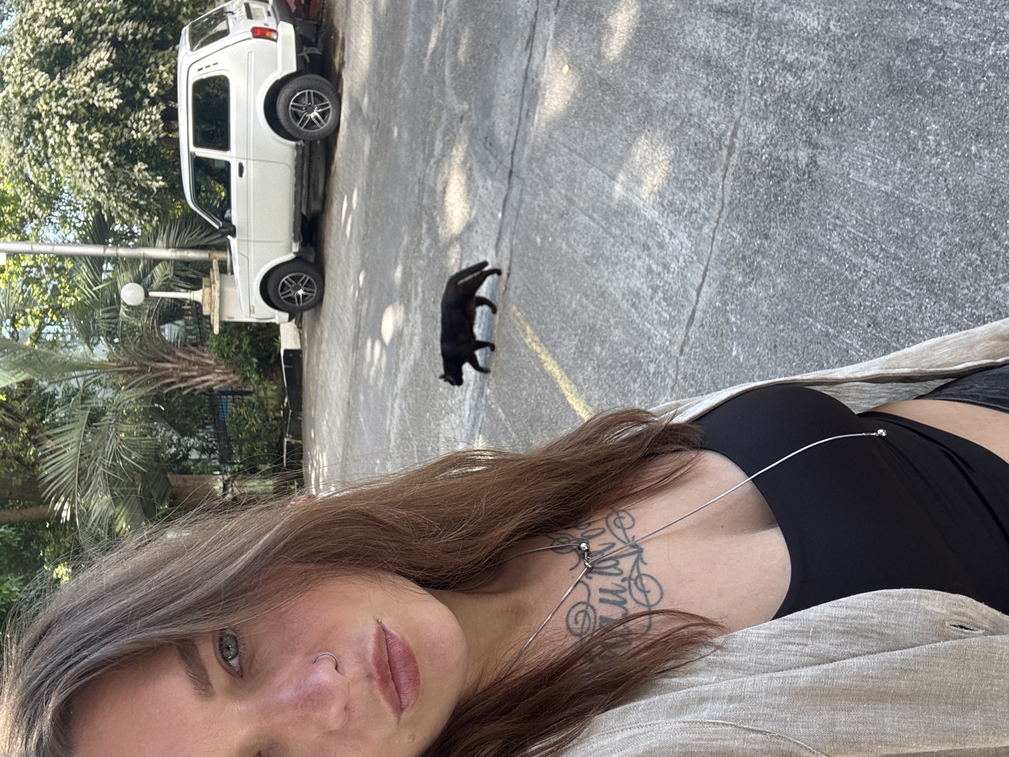 The image depicts a close-up selfie of a woman in a paved outdoor area, identified as being in Sochi, Russia. The woman, positioned on the left side of the frame, has long brown hair, blue-green eyes, a silver hoop nose piercing on her left nostril, and visible freckling. She is wearing a light beige open cardigan over a black top, revealing a detailed black ink tattoo on her chest and a long silver necklace. Her lips are slightly parted. In the middle ground, a black domestic cat walks from left to right across the gray asphalt surface, which has visible cracks and faded yellow markings. In the background, to the right of the cat, a white two-door Lada Niva SUV with dark alloy wheels is parked. Further in the background, lush green vegetation, including palm trees and trees with white blossoms, is visible. A white street lamp on a decorative pillar stands behind the parked vehicle. The scene is illuminated by bright daylight, casting distinct shadows.