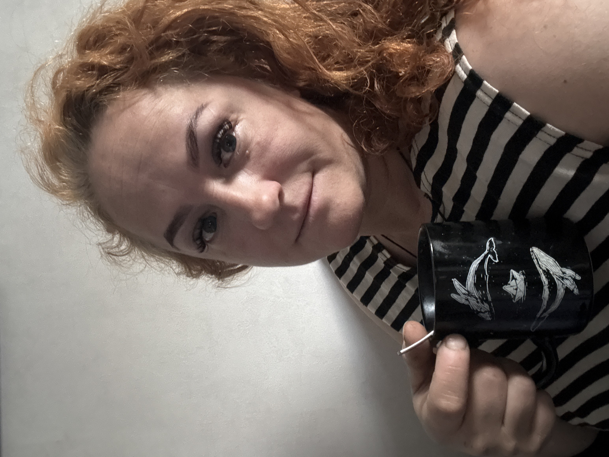 A woman with curly red hair is holding a black mug with an image of whales on it. She is wearing a black and white striped tank top. The background is a plain white wall. There is no evidence to suggest this image was taken in Chaikovsky, Russia.