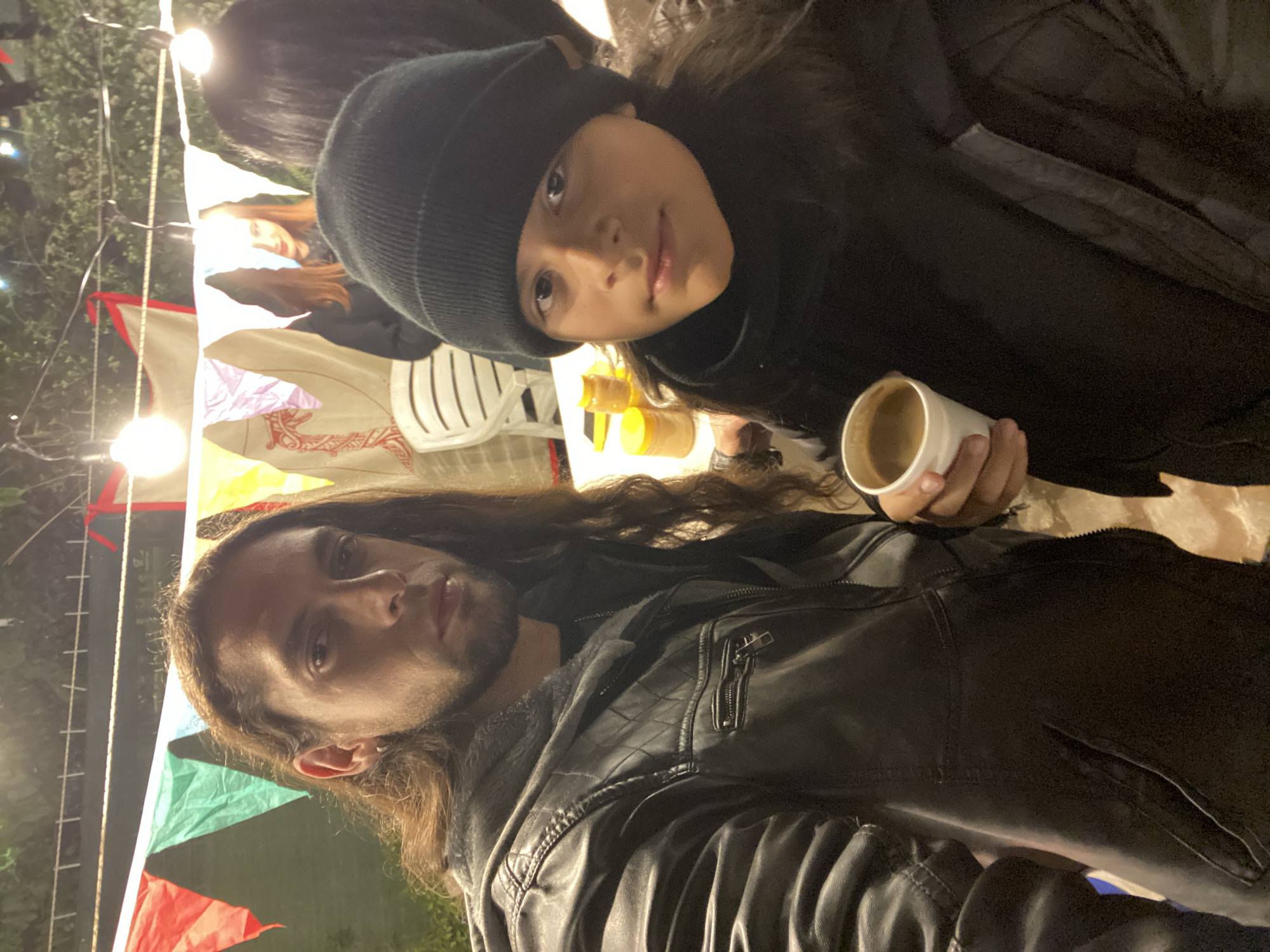 The image depicts two individuals in an outdoor setting, likely at a fair or market.
The person in the foreground has long hair, a beard, and is wearing a black leather jacket. The other person wears a beanie and is holding a cup.
The setting includes colorful triangular flags strung overhead and string lights. There are also chairs, tents and jars visible in the background.
Based on the cultural elements and architecture, the location might be in the city of Buenos Aires, Argentina.